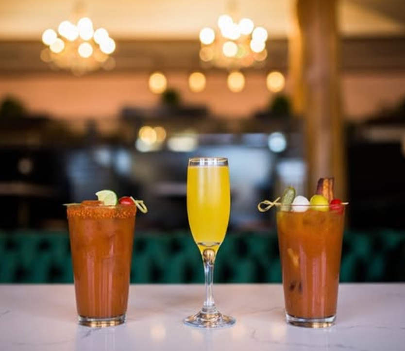 A pink cocktail garnished with a purple edible flower and lavender, served on a wooden tray along with lemon slices and purple and white flowers, on a pink background.