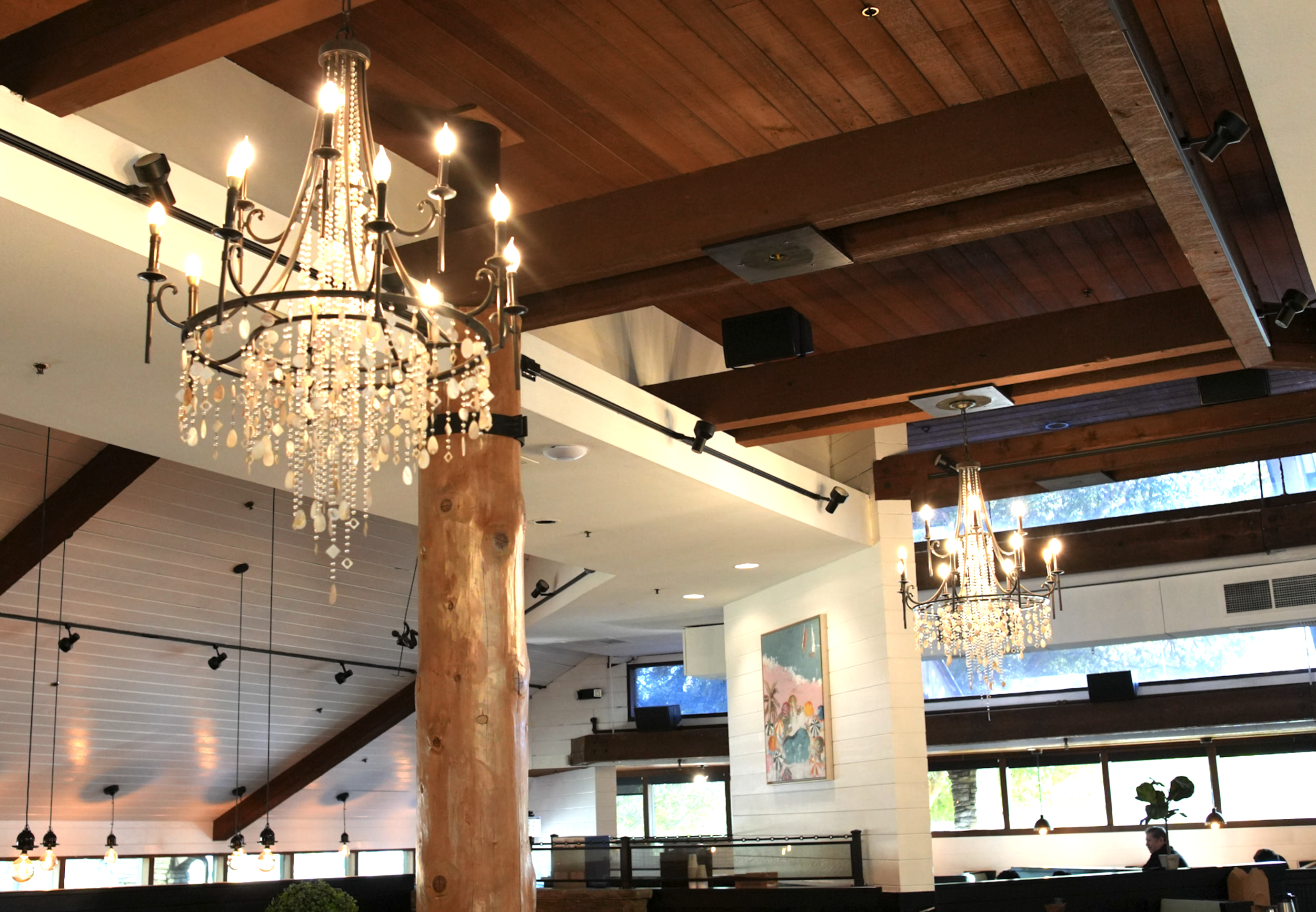 Interior of a restaurant or cafe featuring chandeliers, wooden ceiling beams, and large windows.