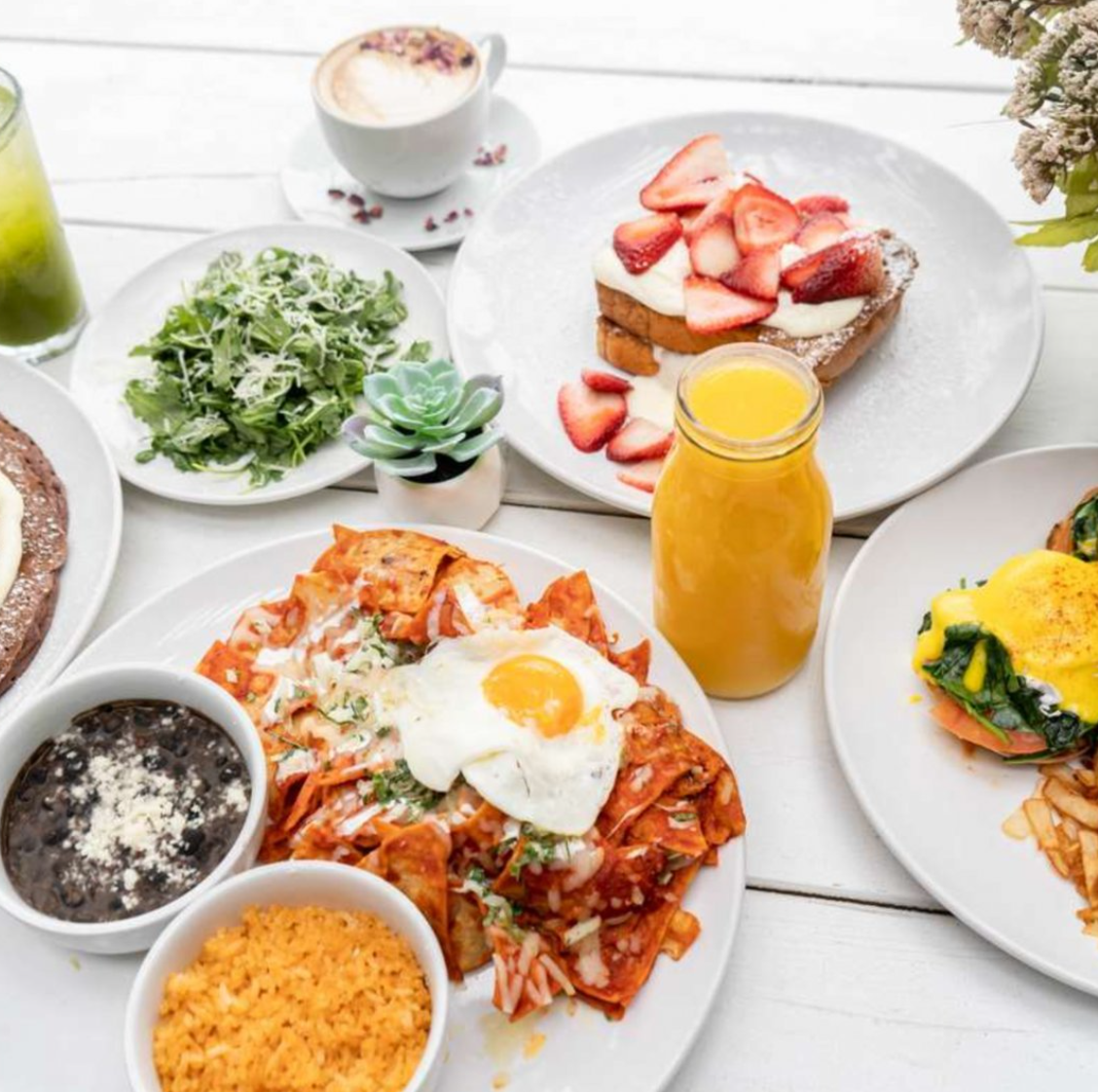 Breakfast spread featuring a plate of huevos rancheros with fried egg, salsa, and rice, a coffee drink with foam, a green juice, a small salad, a strawberry-topped French toast with cream, and a bottle of orange juice on a white table.