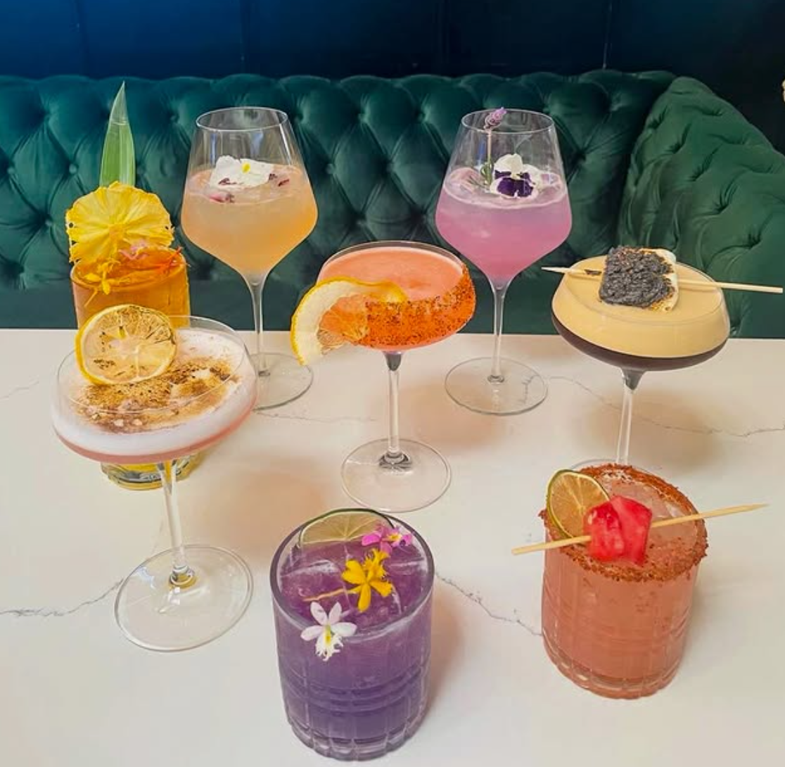 A variety of colorful cocktails on a white marble table with a green tufted booth in the background.