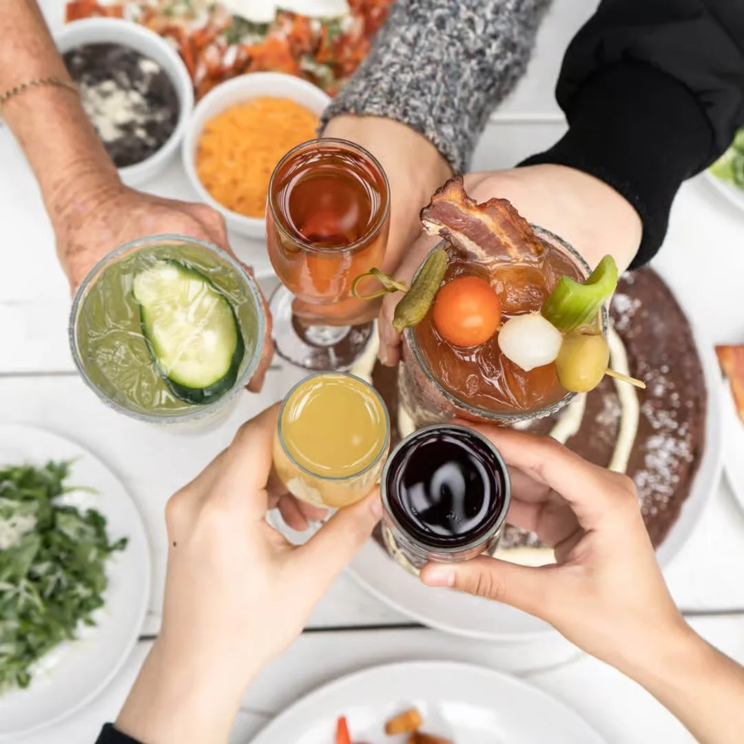 Four people clinking glasses of various beverages over a table with food, including a large bowl of what appears to be a savory dish with bacon and garnishes, and side dishes like a salad and bowls of condiments.