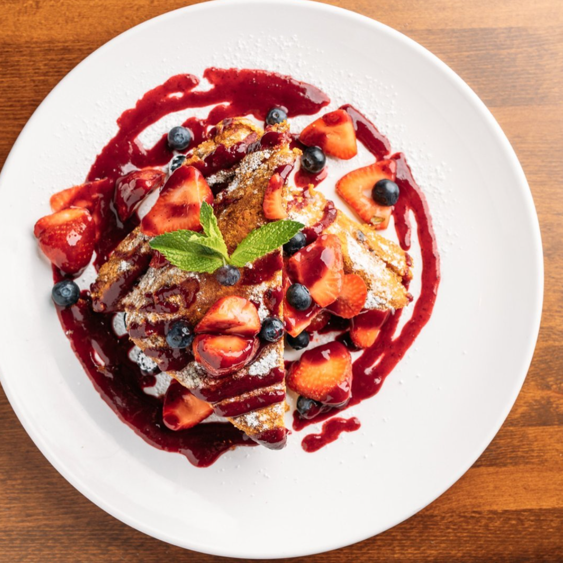 Dessert plate with French toast topped with strawberries, blueberries, powdered sugar, a mint leaf, and berry sauce.