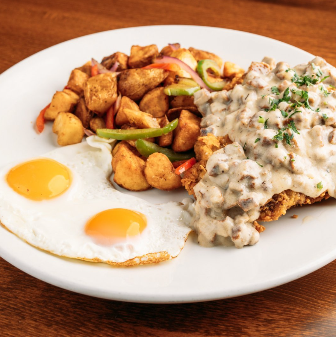 Plate with two sunny-side-up eggs, seasoned roasted potatoes with bell peppers, and a portion of fried chicken covered in white gravy with herbs.