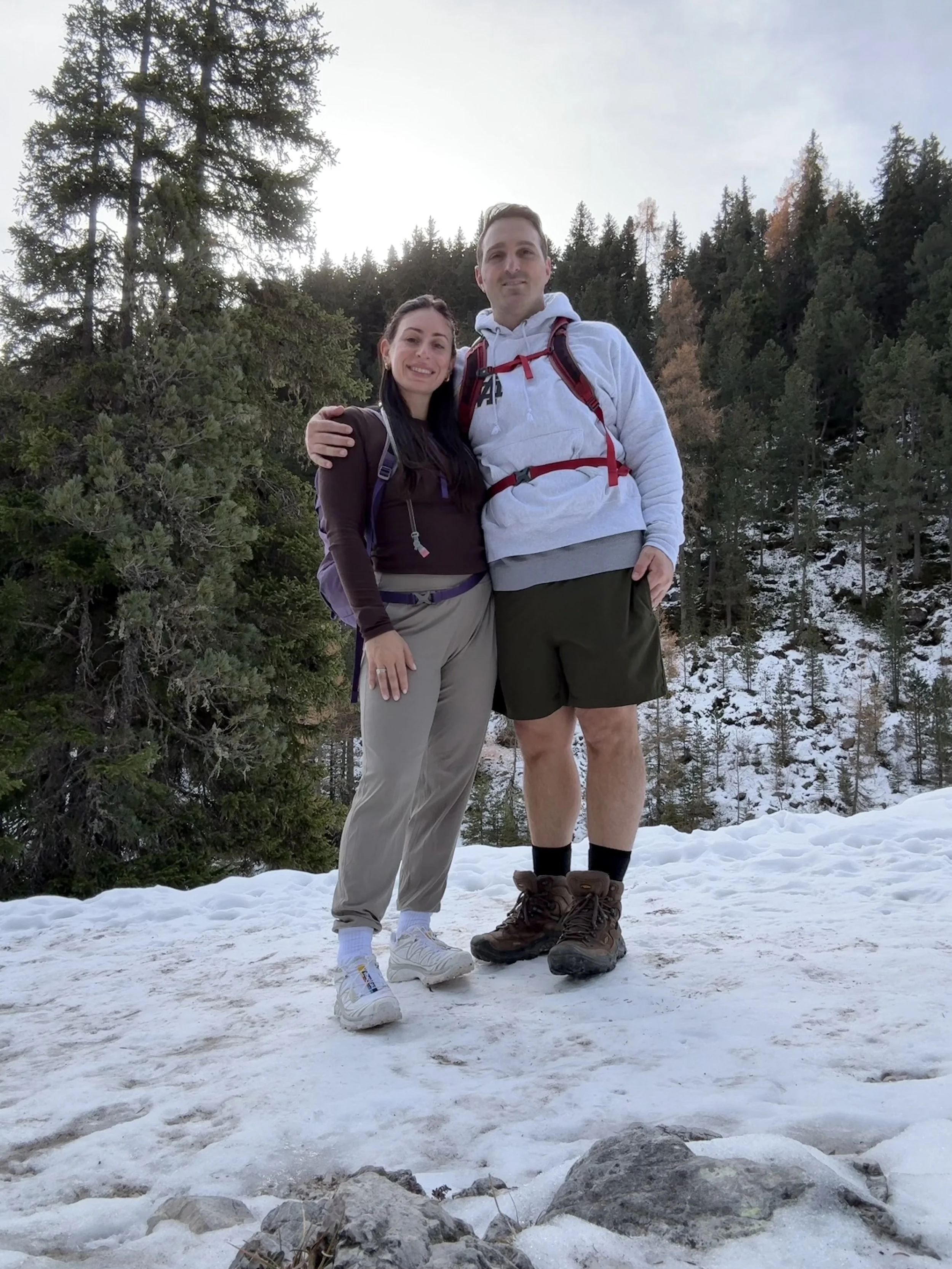 A smiling couple standing on snowy ground in a forested area during daytime, dressed in hiking clothes with backpacks, with tall pine trees and a clear sky in the background.