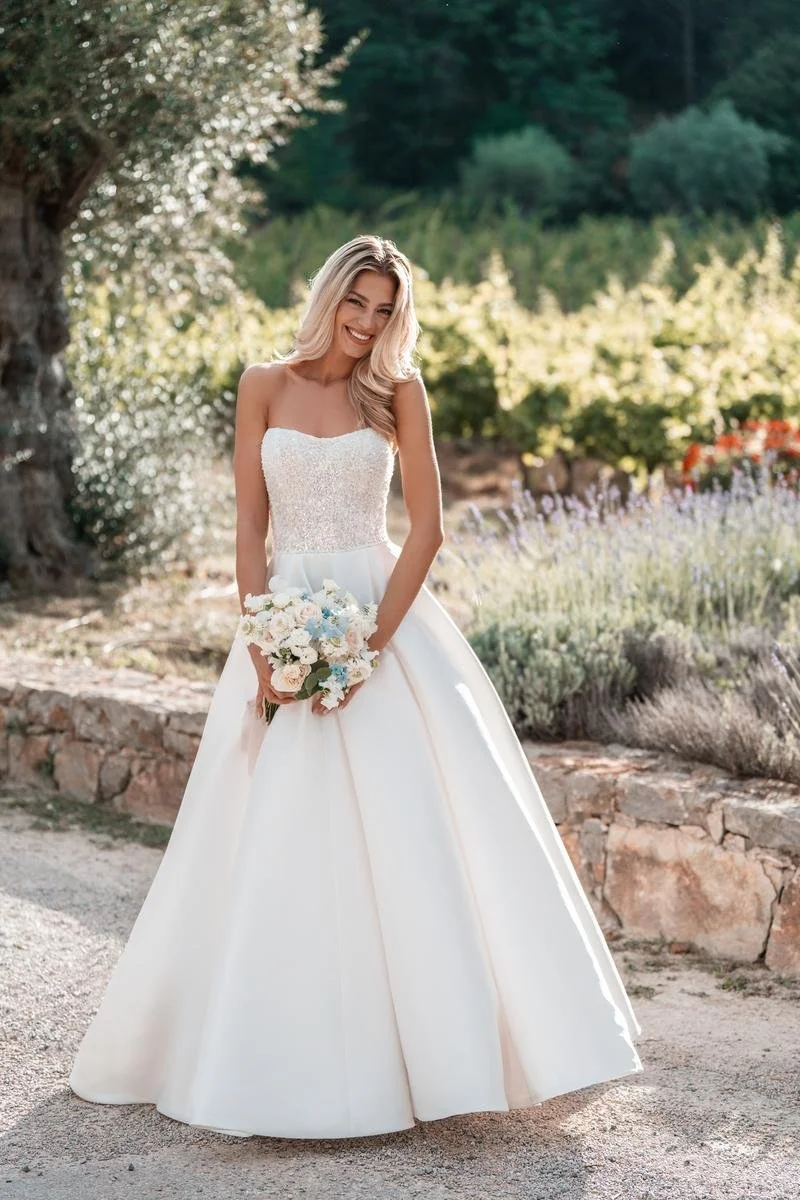 A smiling bride in a strapless white wedding gown holding a bouquet of flowers, standing outdoors in a garden setting with trees and flowers in the background.