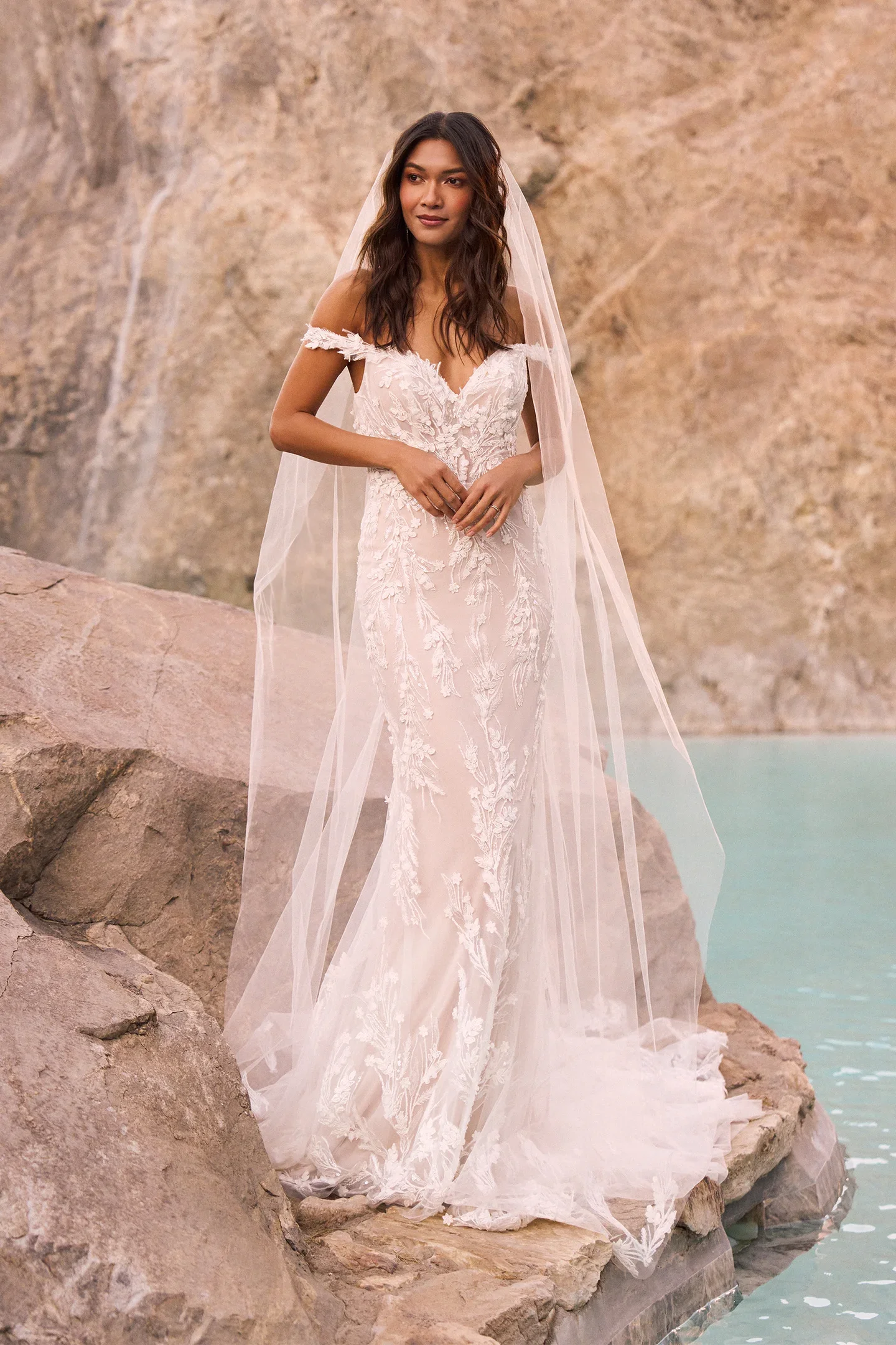 A woman in a white wedding dress stands on a rocky shoreline with water behind her.