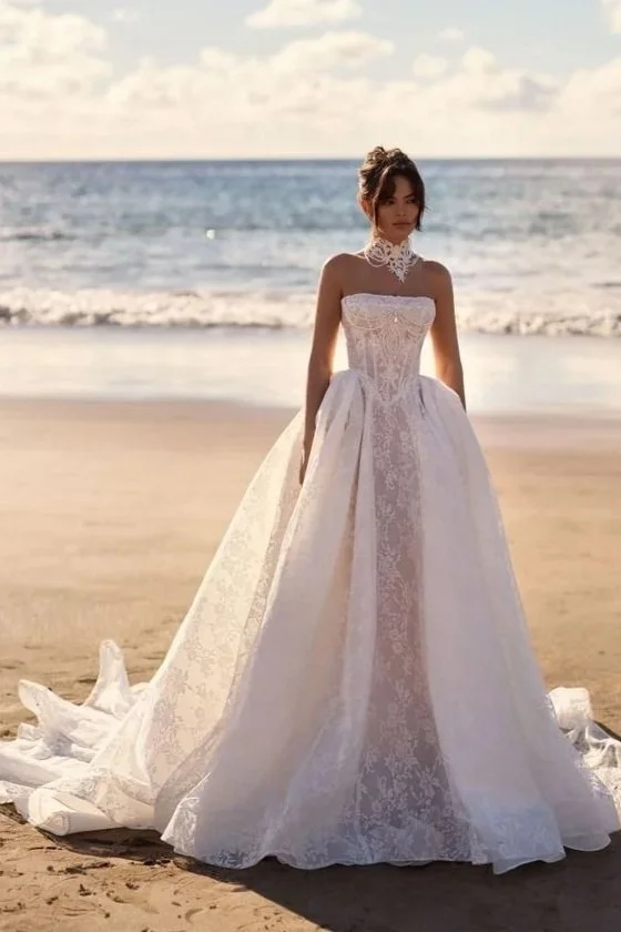 A woman in a white wedding gown standing on a sandy beach with the ocean in the background.