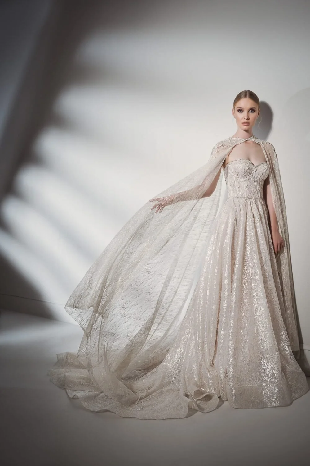 A woman wearing an elegant, light-colored, ornate wedding gown with a matching cape, standing against a plain wall.