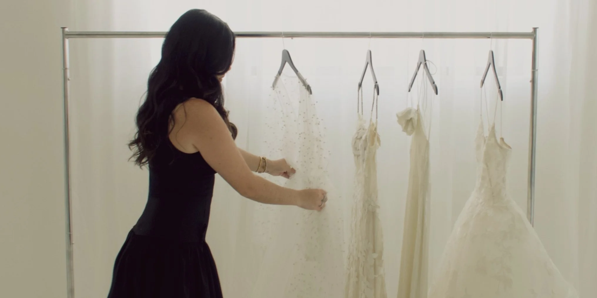 A woman in a black dress is looking at and touching a white dress hanging on a rack, with several other white dresses hanging behind her.