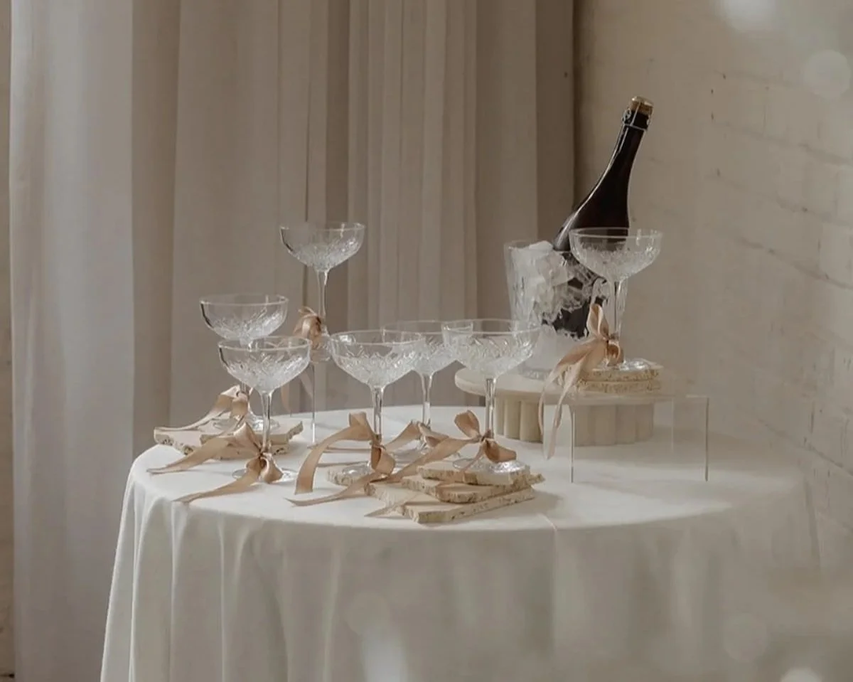 A table with a white tablecloth holding a champagne bottle in ice, eight crystal glasses tied with beige ribbons, and coasters or small plates. The background has light-colored curtains and white brick wall.