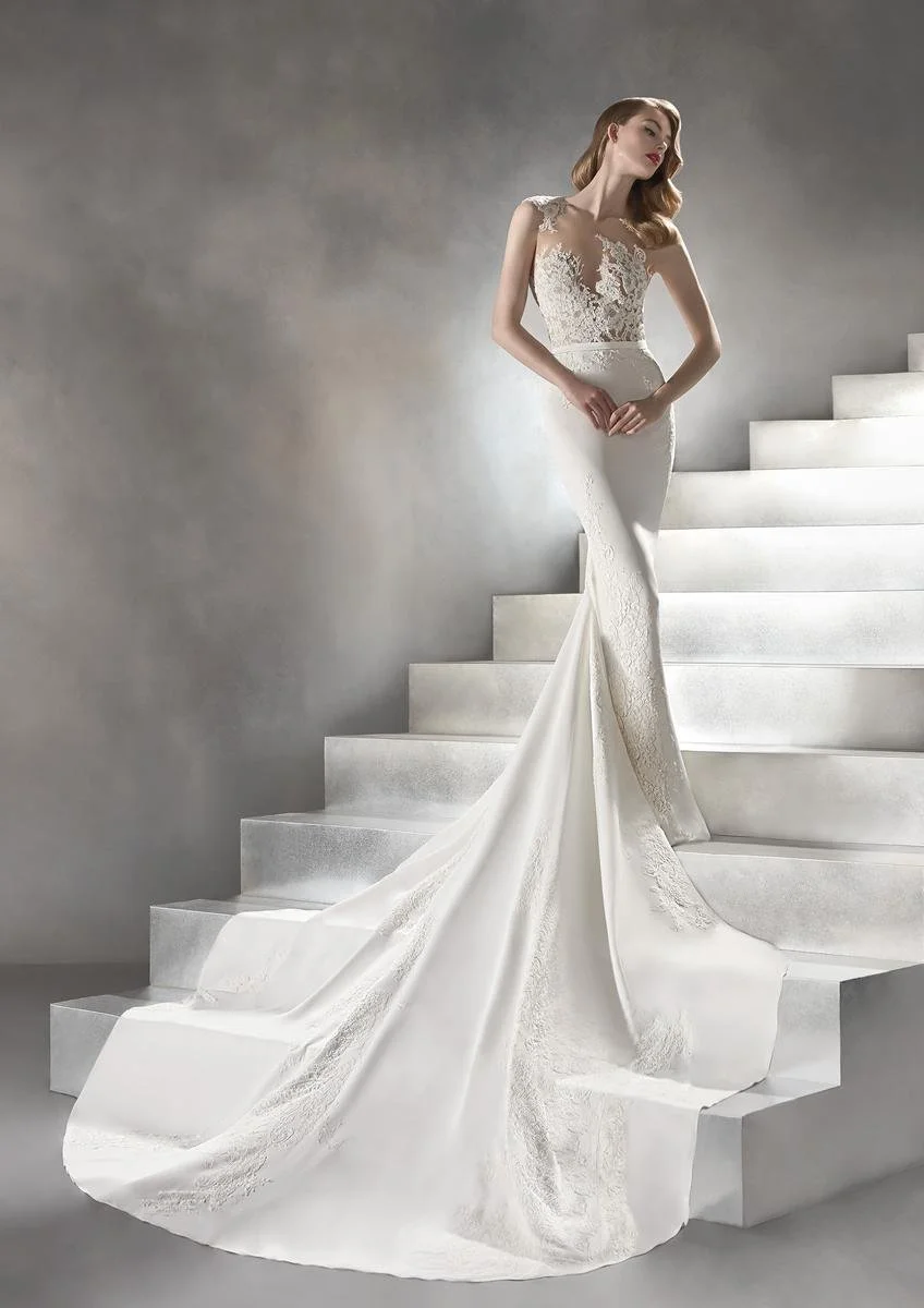 A woman in a white wedding dress with lace details, standing on white stairs against a gray background.