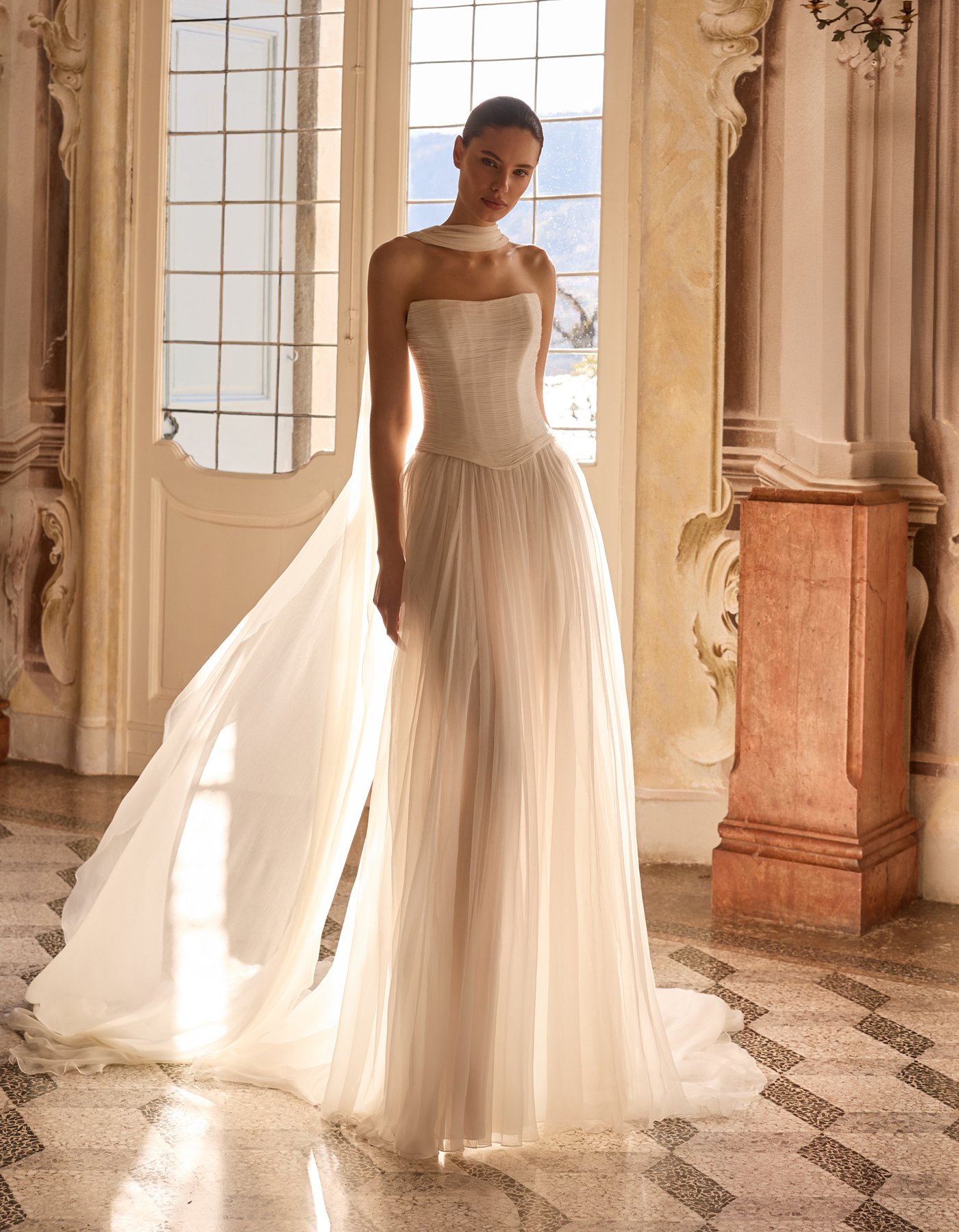 Woman in a strapless white gown with a train standing indoors near a large window with sunlight streaming in, ornate walls, and a marble pedestal.