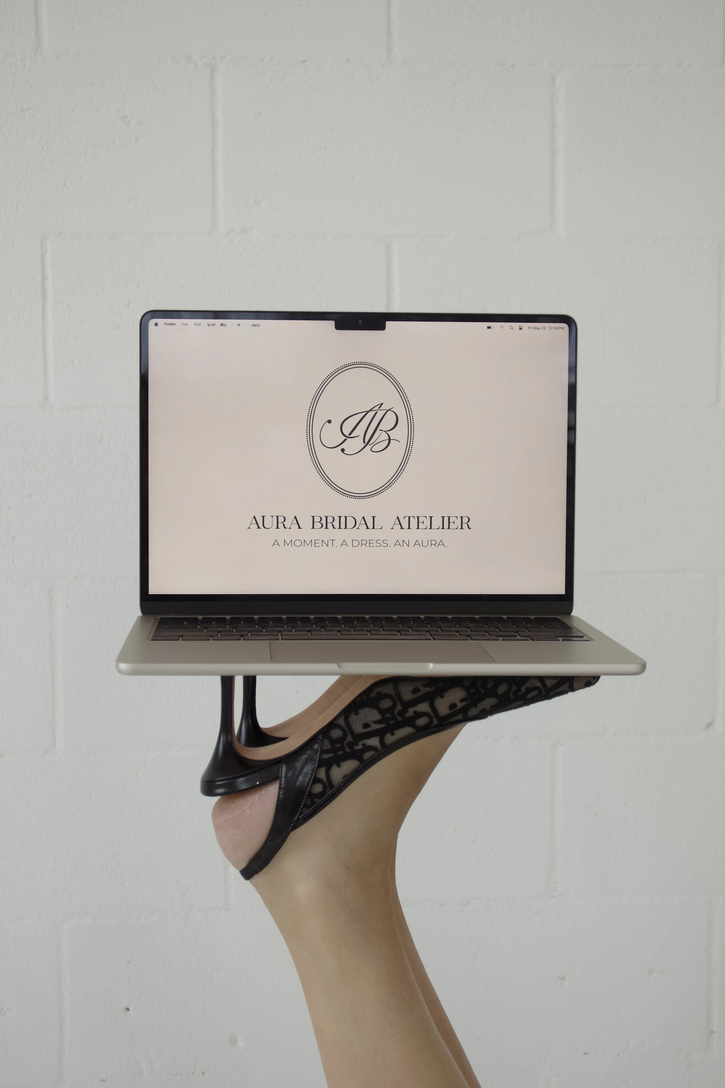 A person holding a laptop displaying the logo for Aura Bridal Atelier on a white wall background.