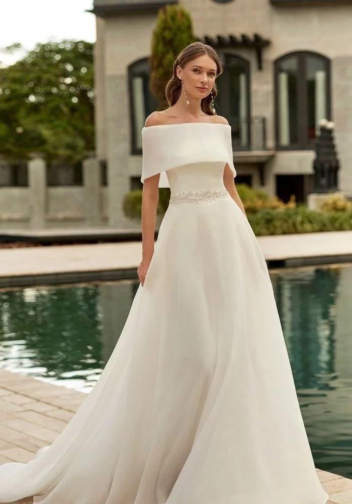 A woman in an elegant off-shoulder white wedding gown standing outdoors near a pool in front of a house.