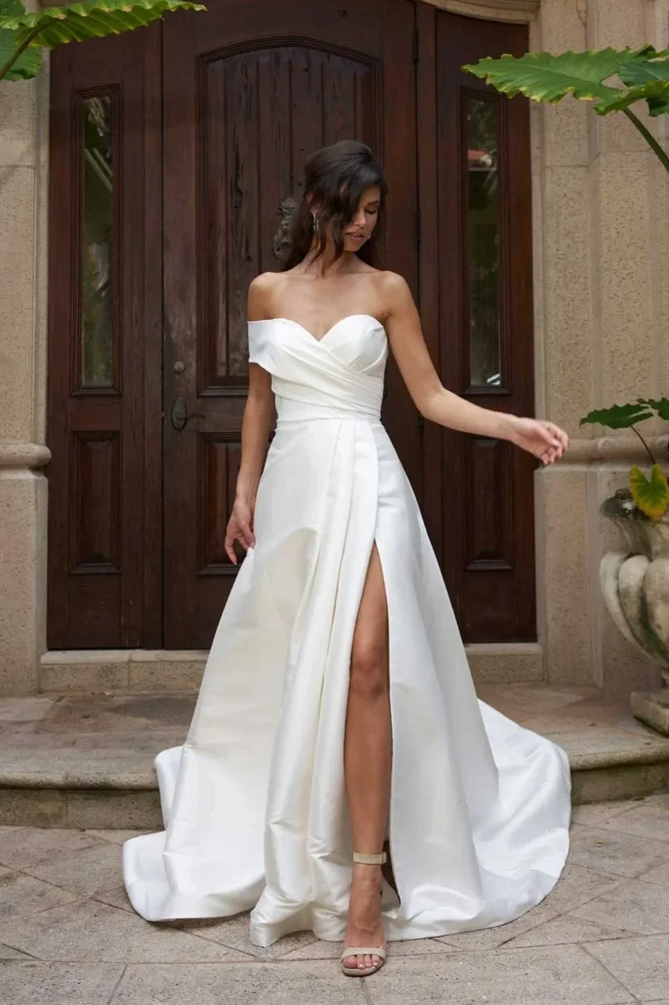 A woman in a white off-the-shoulder wedding dress with a high slit, standing on a stone porch in front of a wooden door, surrounded by plants.