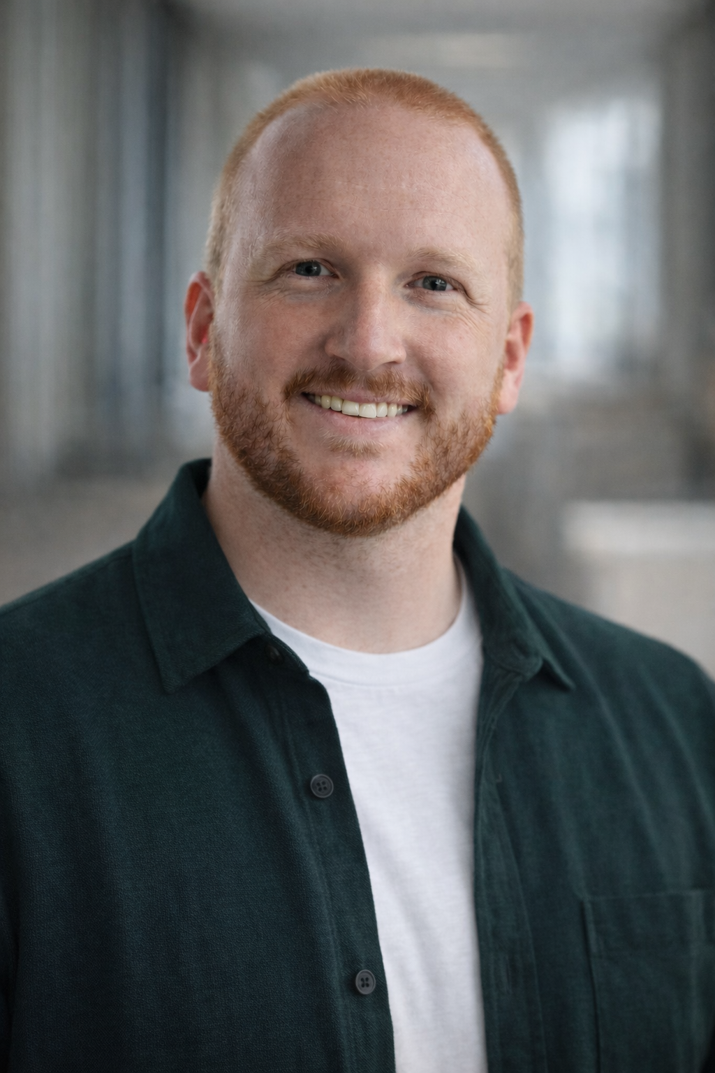 A smiling young man with short red hair and a beard, wearing a dark green jacket over a white T-shirt, standing indoors with a blurred metallic background.