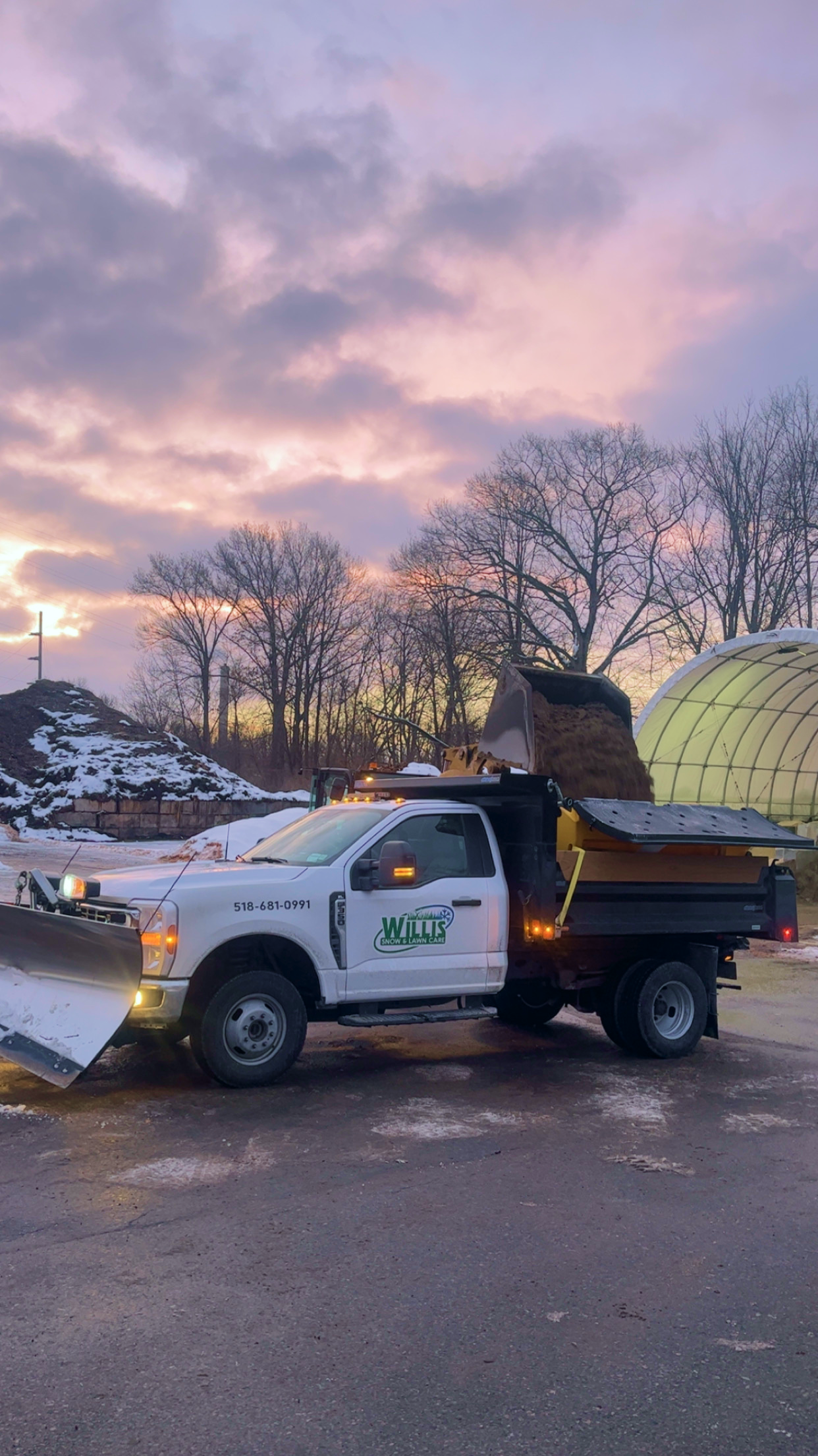 A snow plow truck equipped with a front blade and a large plow on the back, parked on a snowy road during sunset with bare trees.