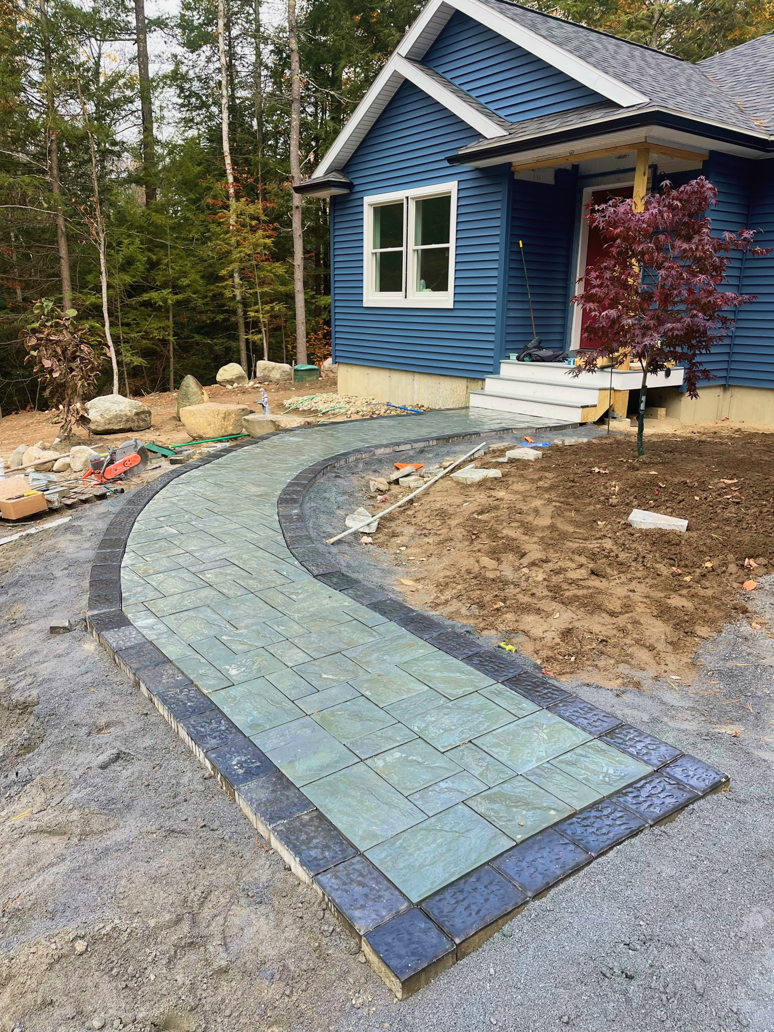 Curved stone pathway under construction in front of a blue house with white trim and a small porch. The yard around the pathway is being landscaped, with dirt and rocks, and a small purple-leafed tree planted near the house. Construction tools and supplies are visible along the pathway.