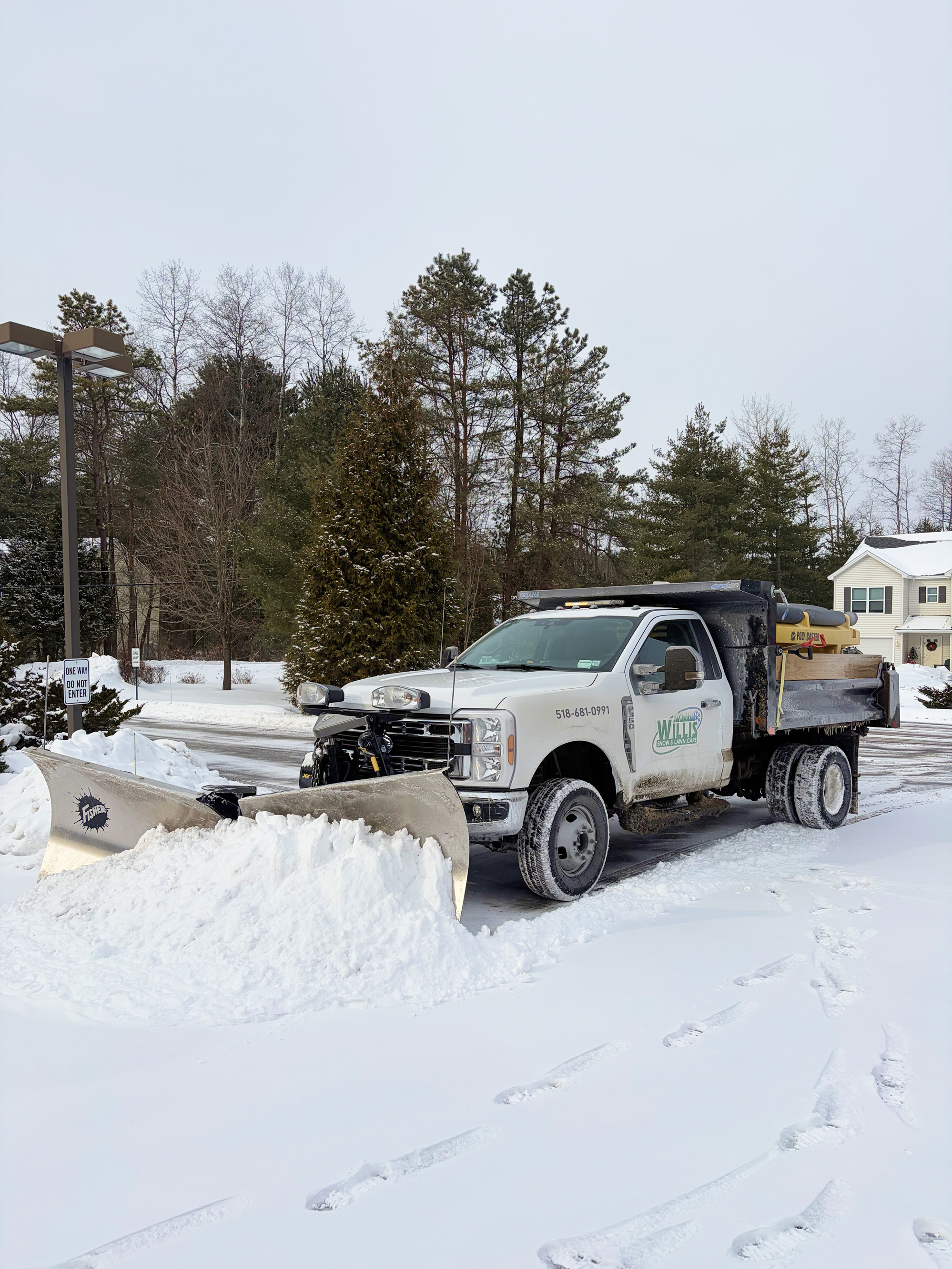 A white snow plow truck clearing snow from a parking lot during winter, with trees and a house in the background.