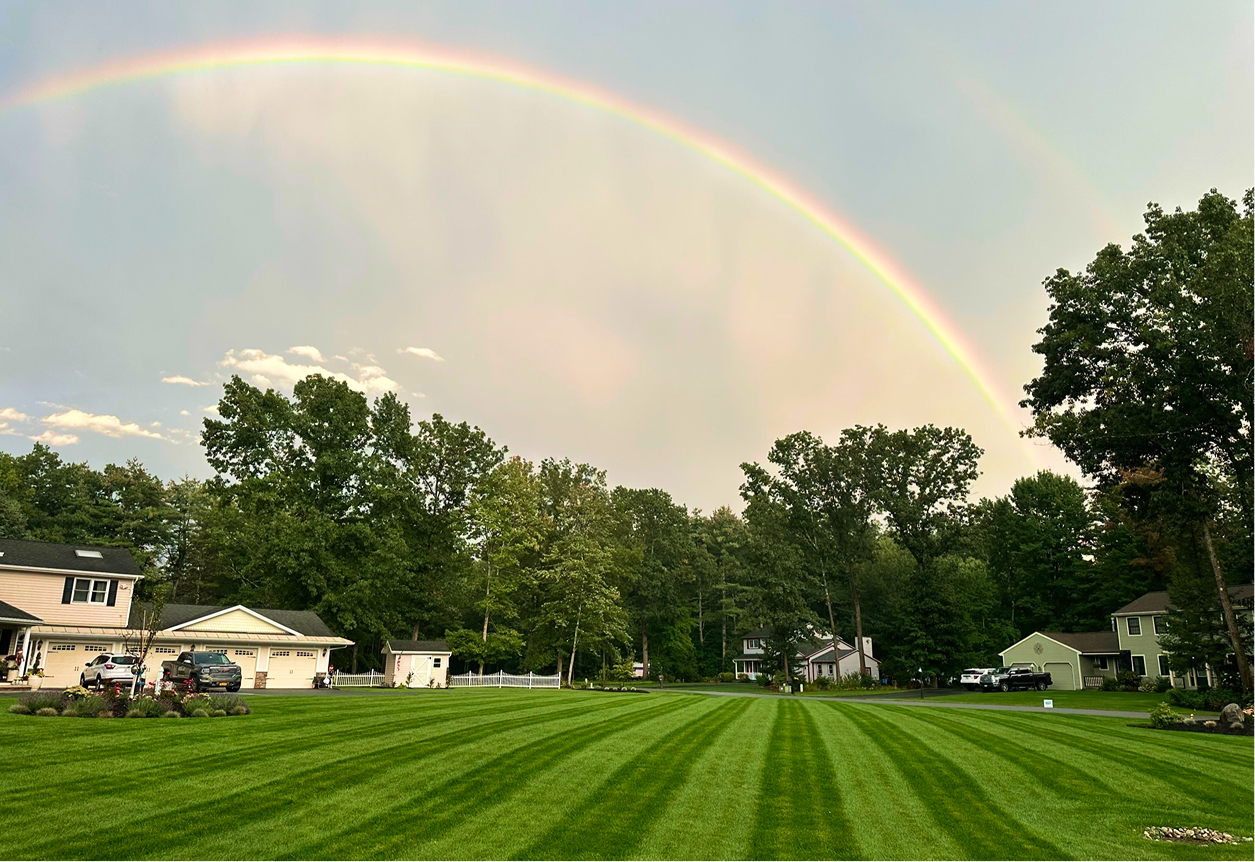 A rainbow arching over a suburban neighborhood with neatly mowed grass and houses.
