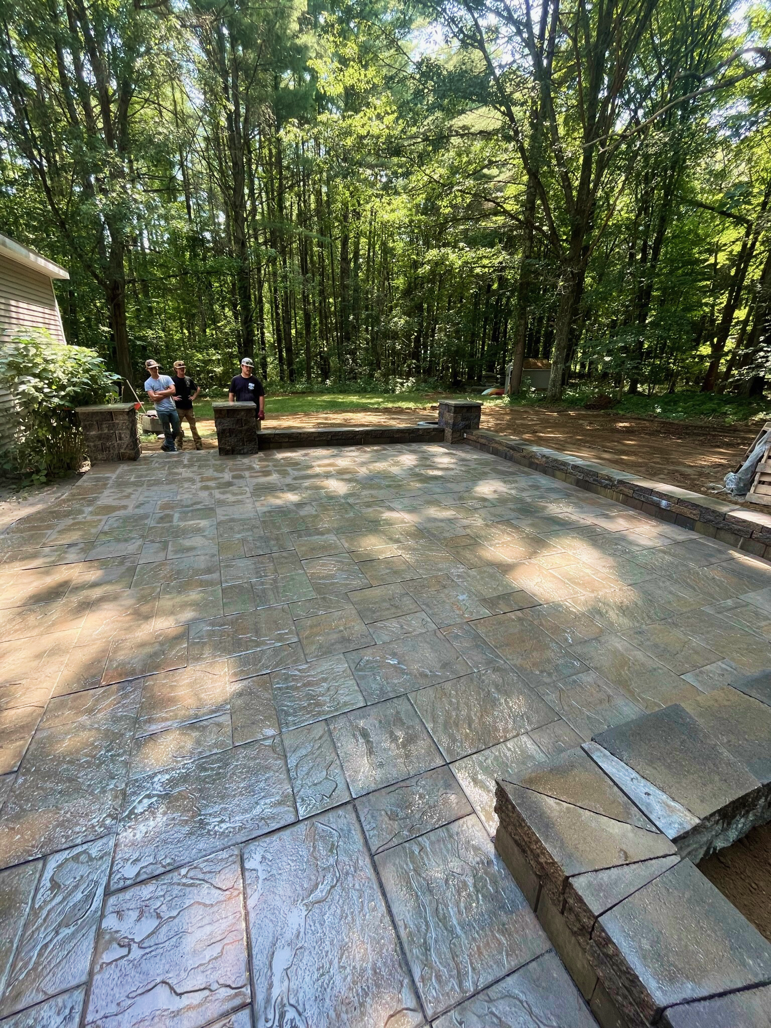 Backyard with new stone patio under tall trees, three people standing and talking near the edge of the patio, sunlight filtering through the leaves.