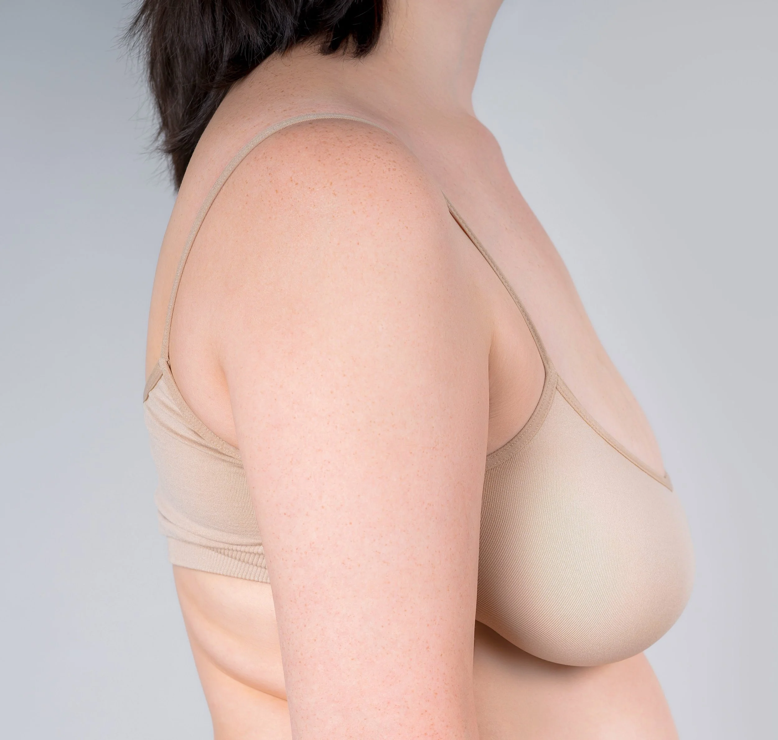 Close-up of a woman wearing a beige bra, showing her shoulder, upper arm, and partial chest against a plain gray background.