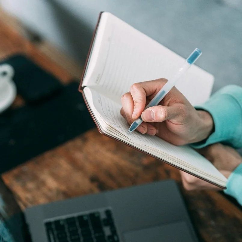 Person writing in a notebook with a pen, with a laptop on a wooden table.