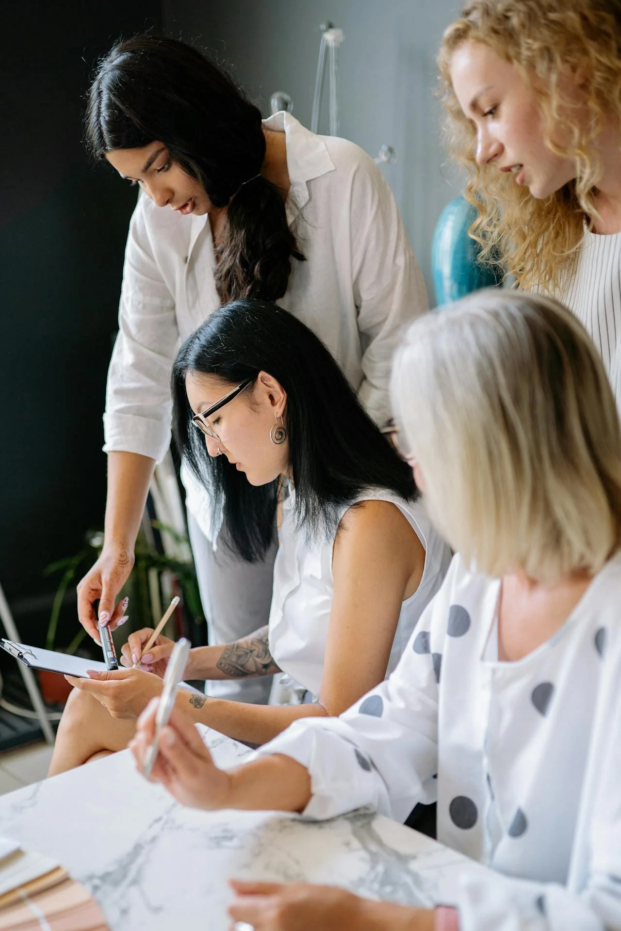 Group of women working together at a table, one writing on a notepad while another points at a tablet, in a collaborative work environment.