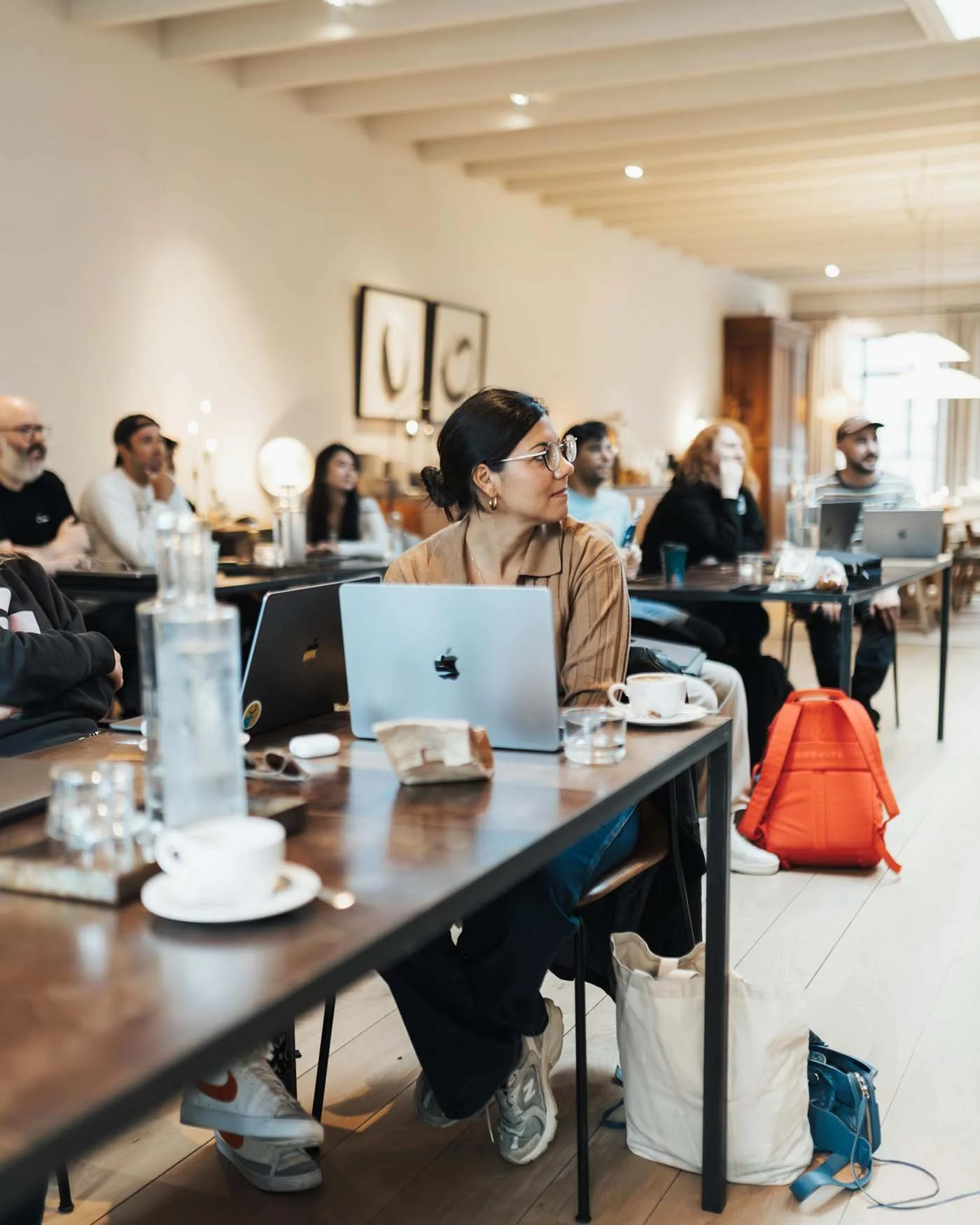 A group of people attending a workshop or seminar in a well-lit room, sitting at tables with laptops, drinks, and snacks.