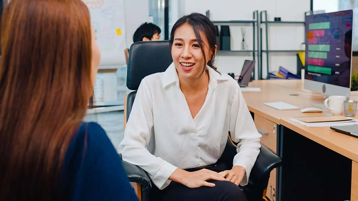 Two women talking in an office, one smiling and facing the camera, the other with her back to the camera.