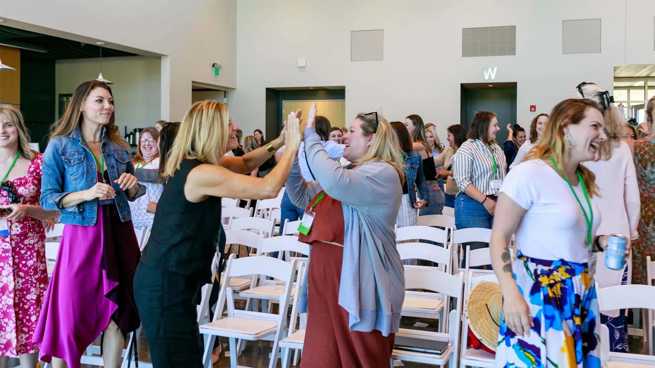 Group of women at a conference or seminar, engaging and high-fiving each other, with chairs and a spacious indoor venue background.
