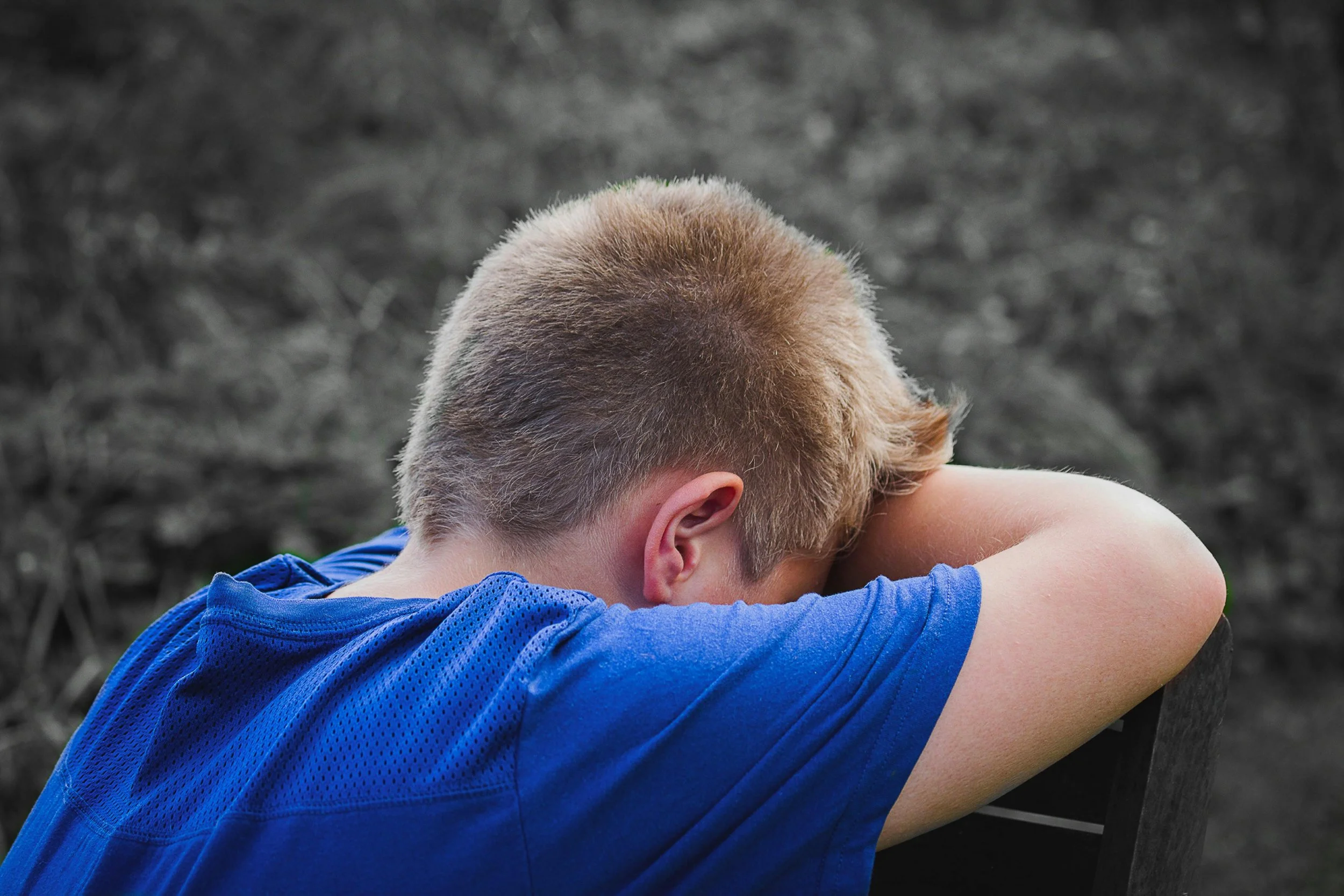 A young person with short blond hair is sitting on a bench, with their head resting on their arms, facing downward, wearing a blue shirt. The background appears to be a blurred outdoor setting.