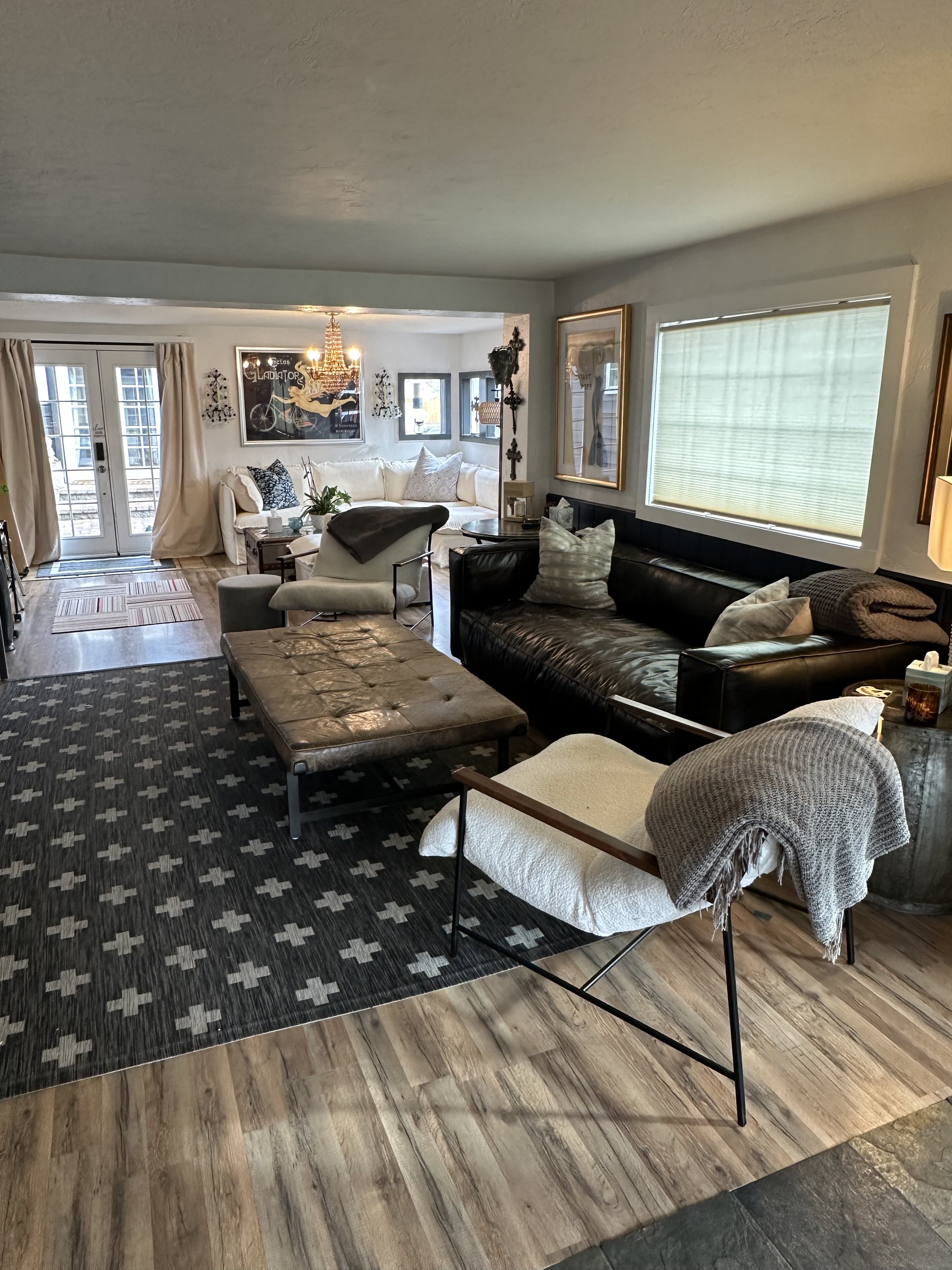 A cozy living room with a black leather sofa, a white armchair with a blanket, a tufted ottoman, and a patterned area rug on wooden floors. Glass doors lead outside, and a plush white sectional sofa is in the background. Decor includes wall art, a ch