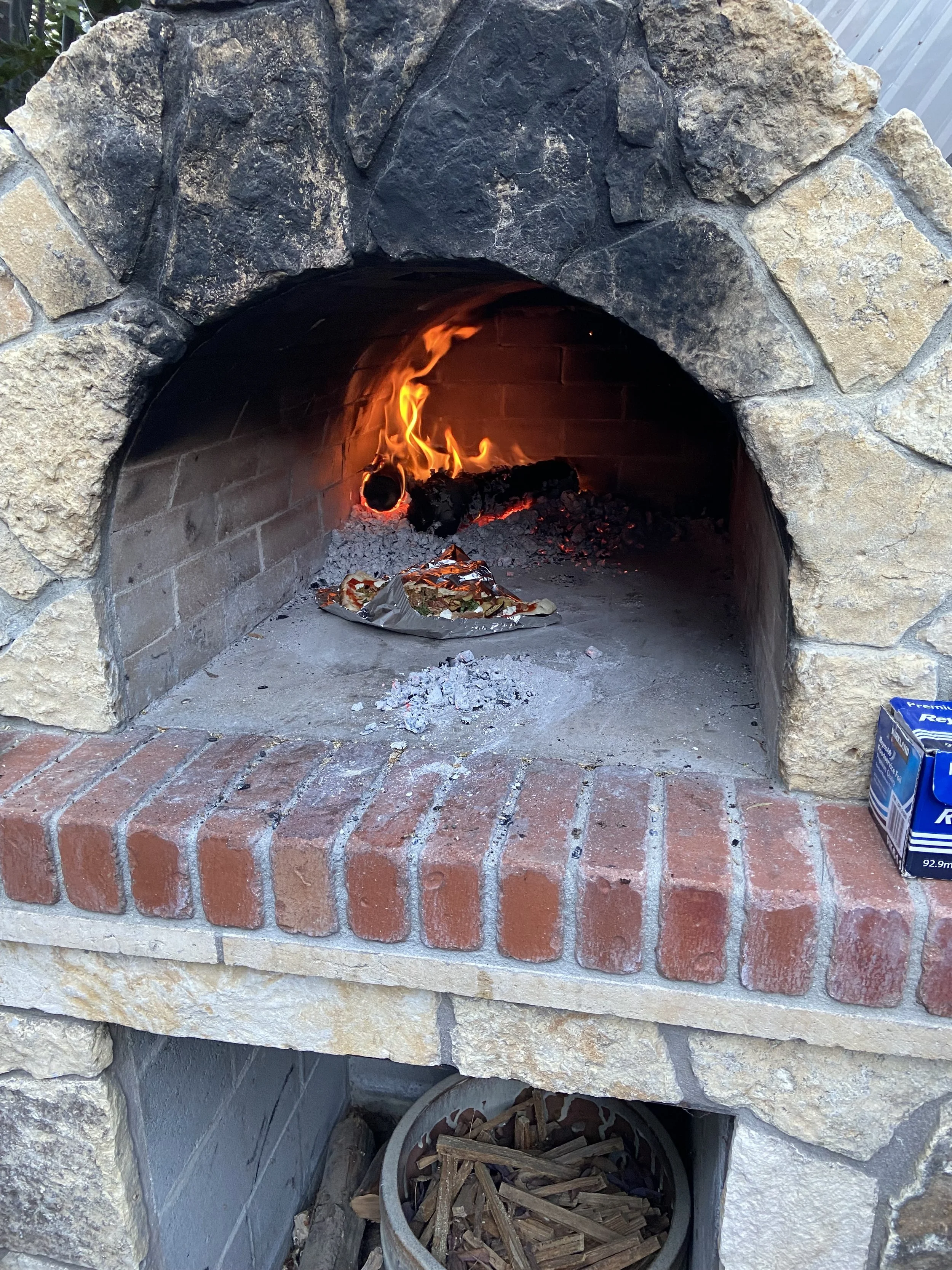 Outdoor brick and stone fireplace with a lit fire inside, flames visible, and a paper pizza box on the hearth. There is a box of Reynolds Wrap foil on the right side of the hearth and a bucket of kindling or wood at the base of the fireplace.