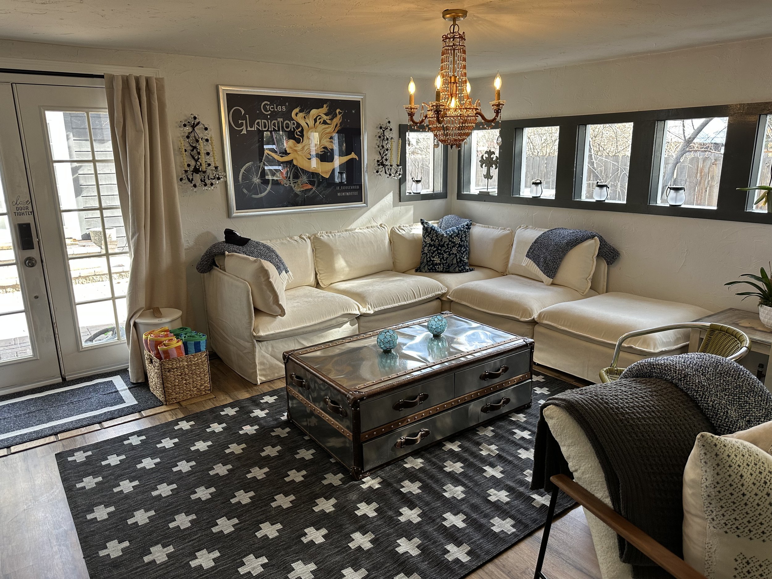 Living room with cream sectional sofa, dark patterned pillows, a glass-top coffee table with decorative spheres, a black-and-white patterned rug, a chandelier, and a wall art poster.