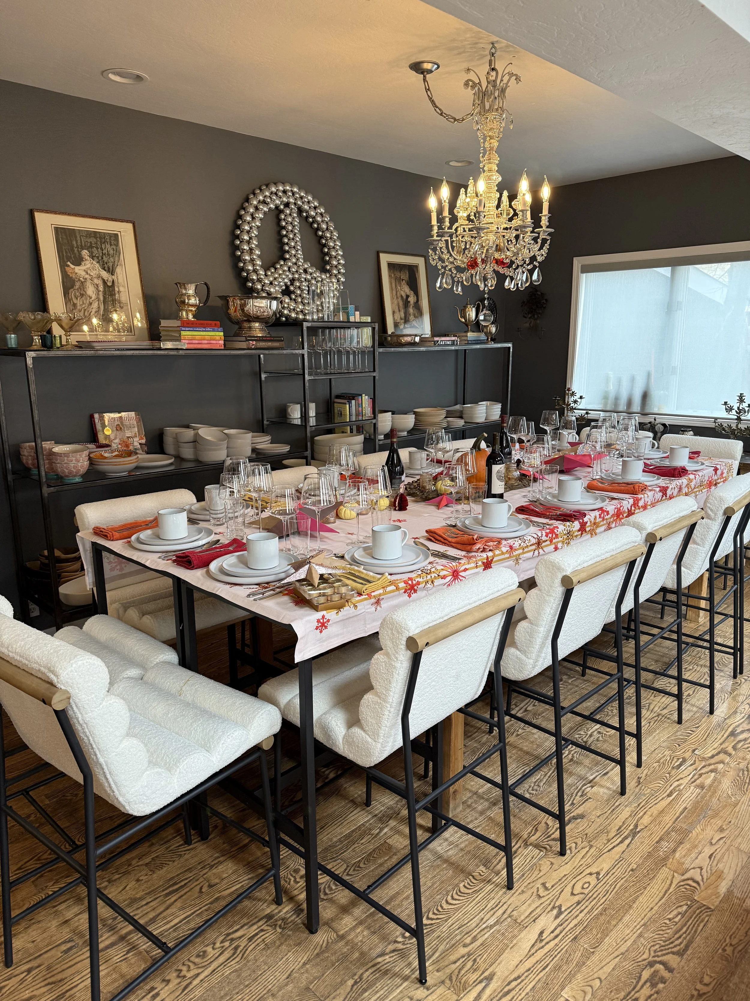 A dining table set for a holiday meal, with white chairs, plates, cups, glasses, and festive decorations, in a room with a gray wall and a chandelier.