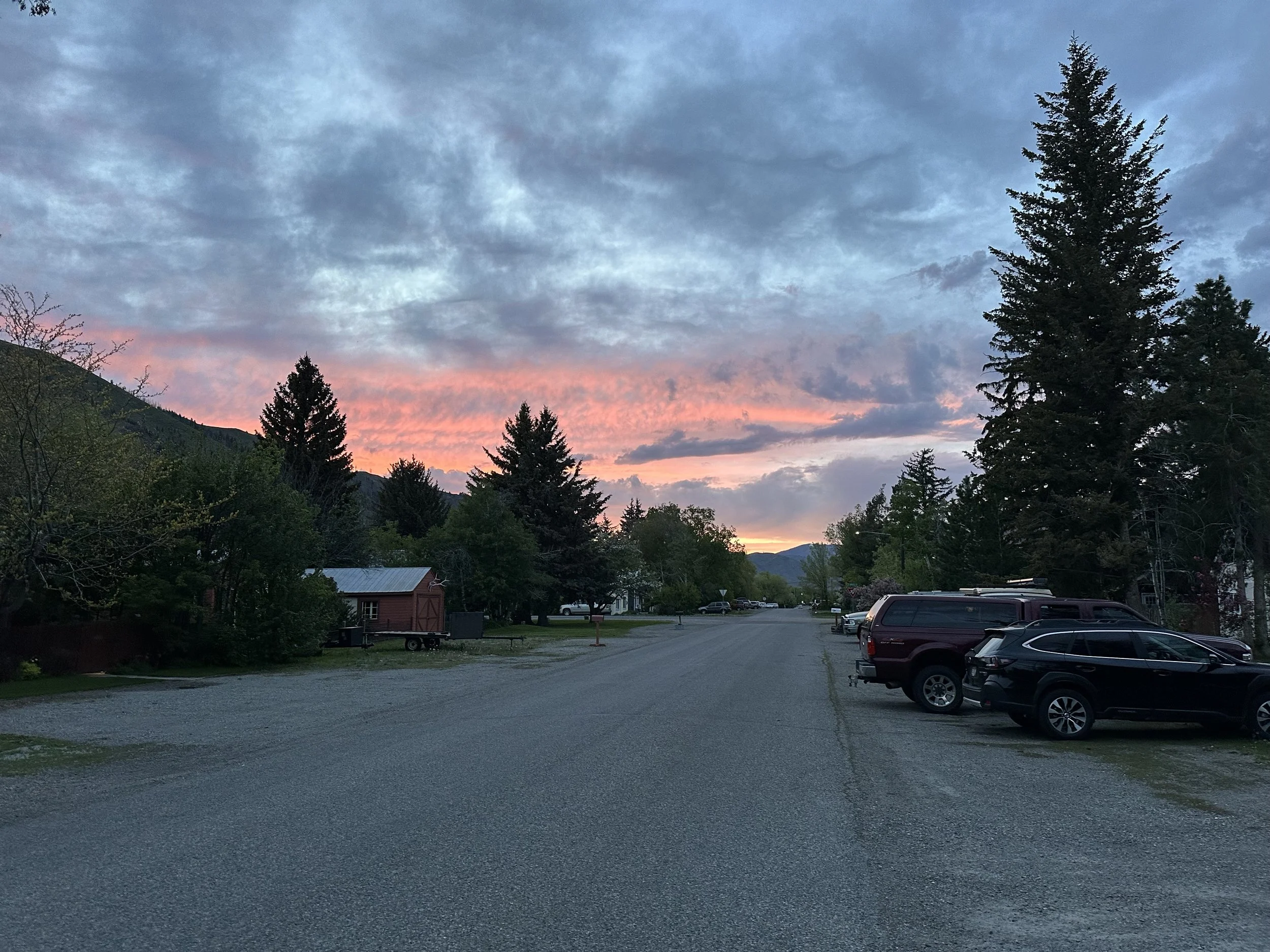 A peaceful outdoor scene of a gravel parking area at sunset, with a backdrop of mountains and a sky painted with soft pink and blue clouds. Tall evergreen trees and small buildings are visible on either side of the lot.