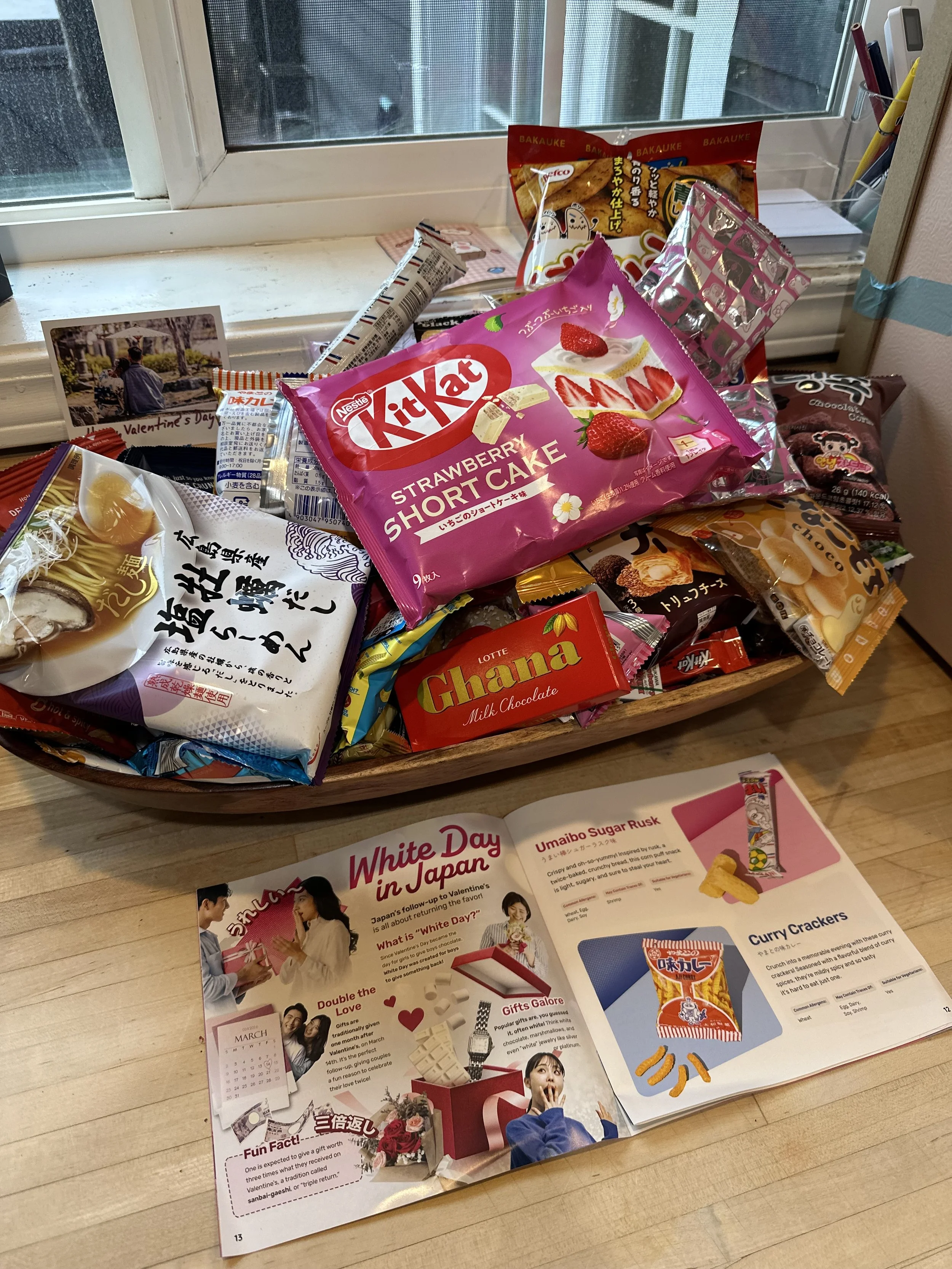 A wooden table with a basket filled with various Japanese snacks, including Kit Kat strawberry shortcake, Chana milk chocolate, and other treats. An open brochure about White Day in Japan lies on the table, featuring images and descriptions of tradit