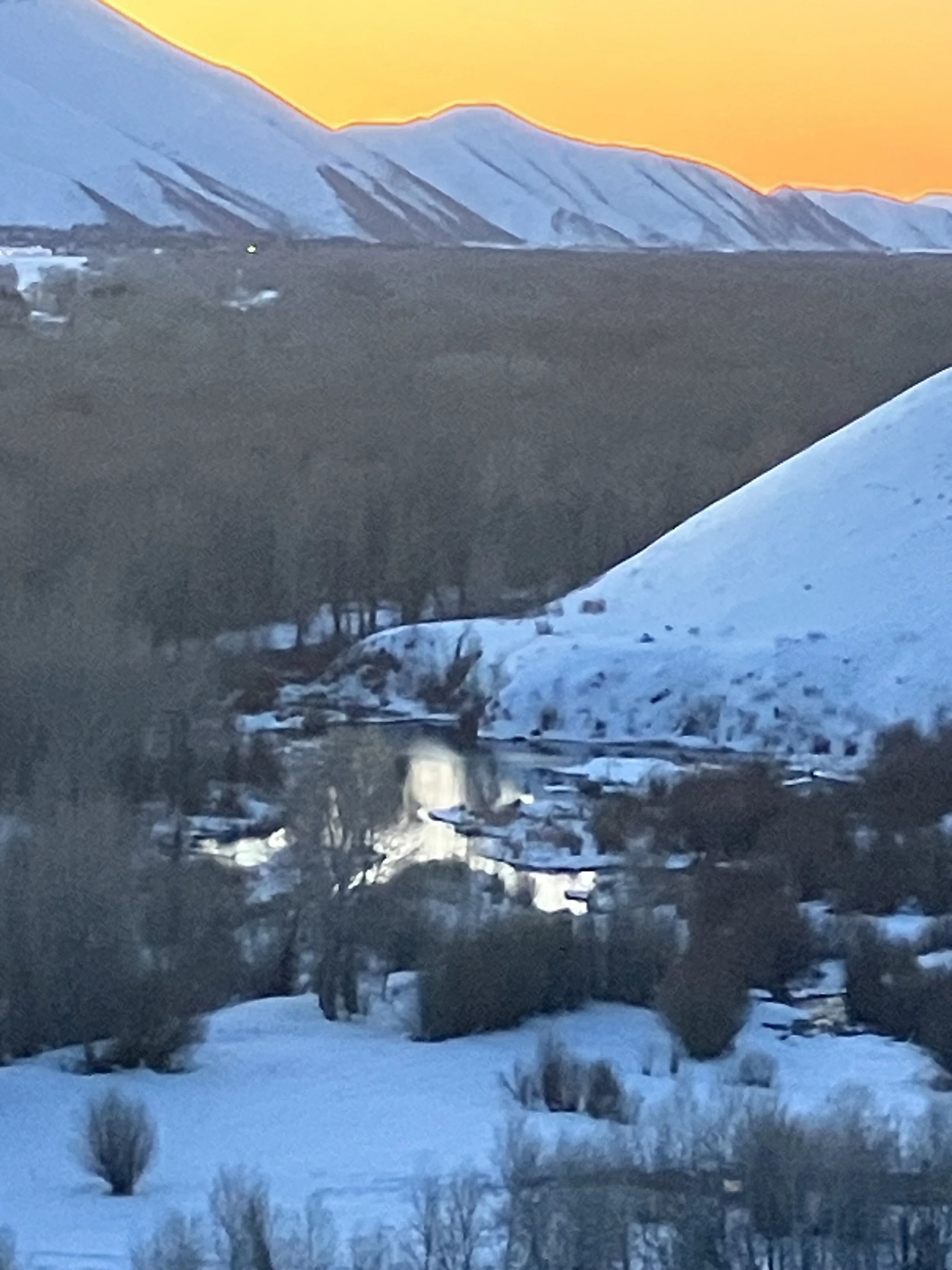 Snow-covered mountain range at sunset with a river and winter trees in the foreground.