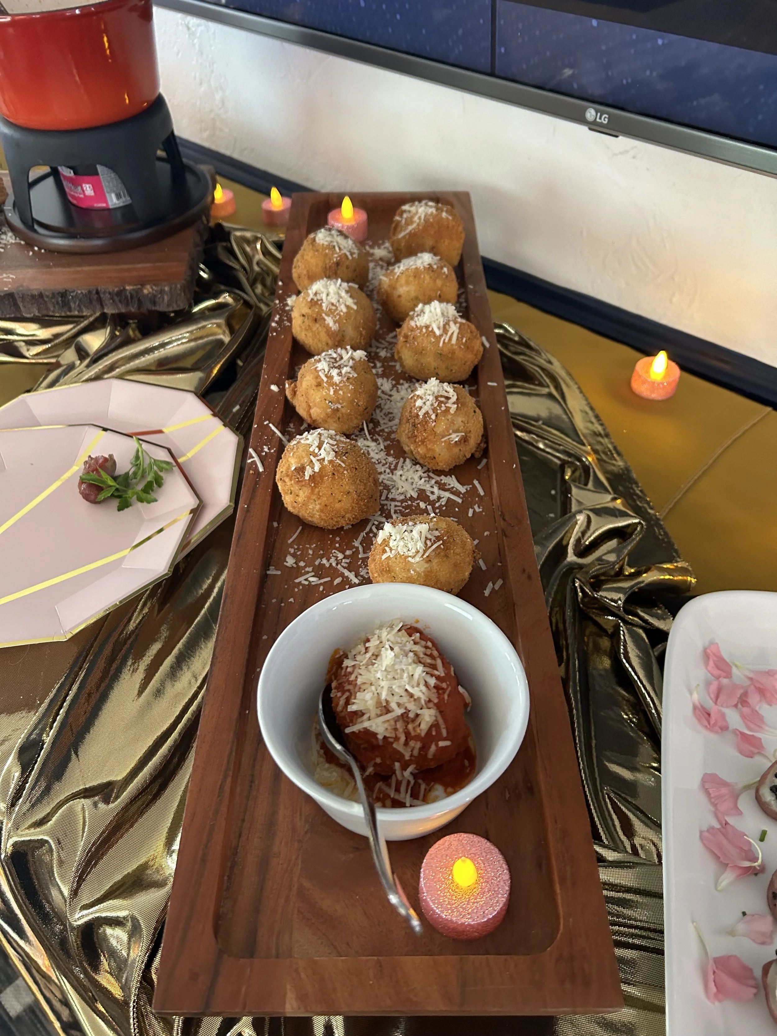 Fried cheese balls sprinkled with shredded cheese on a wooden serving board, with a bowl of marinara sauce, set on a table decorated with gold and pink accents and small pink candle lights.