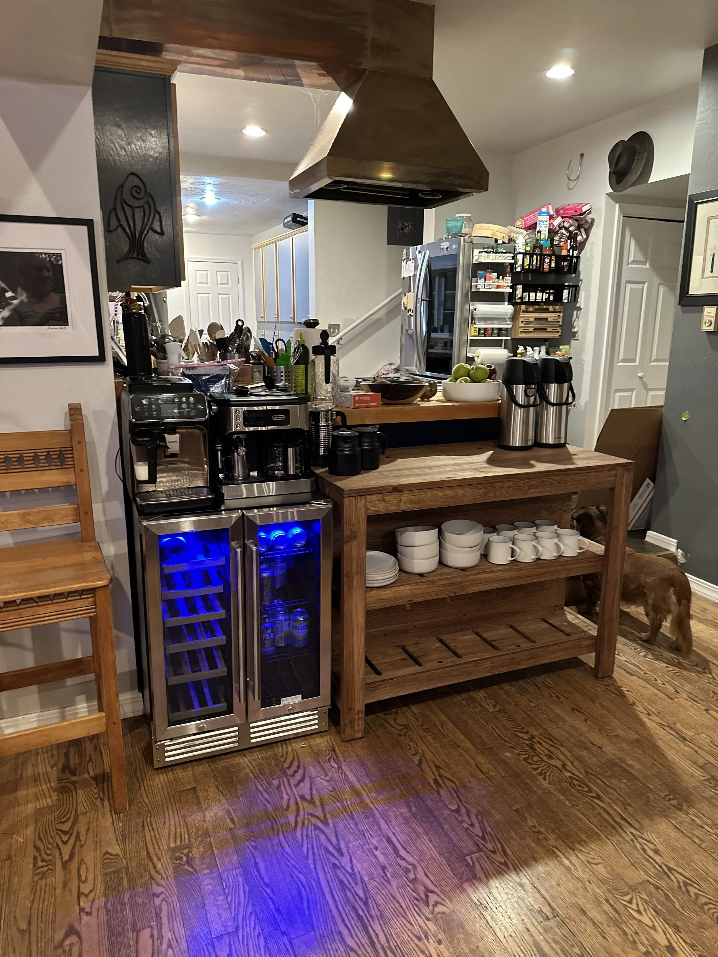 A kitchen corner with a wooden table, white mugs and bowls, silver beverage dispensers, and a small wine cooler, with a dog partially visible behind the table.