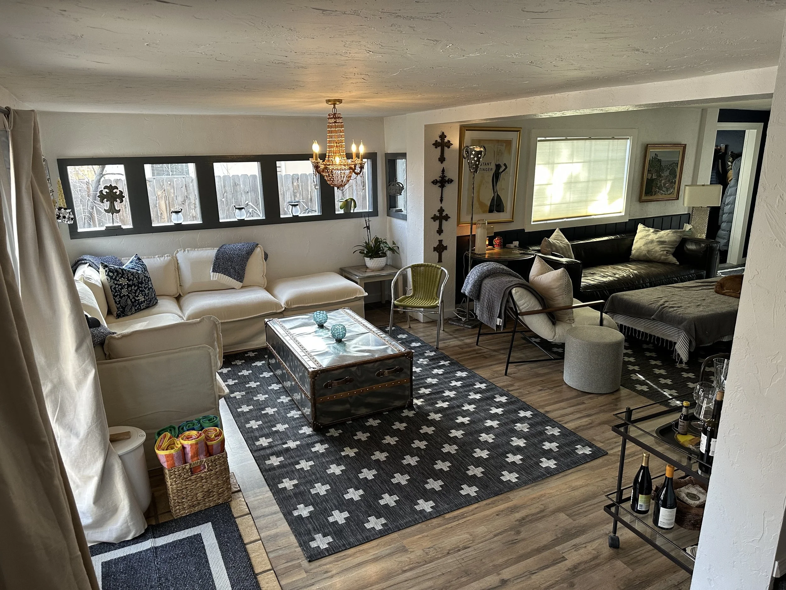 Living room with white sectional sofa, black leather sofa, area rug with white crosses, glass-top trunk coffee table, yellow accent chair, and various decor items including chandelier, pillows, artwork, and decorative crosses.