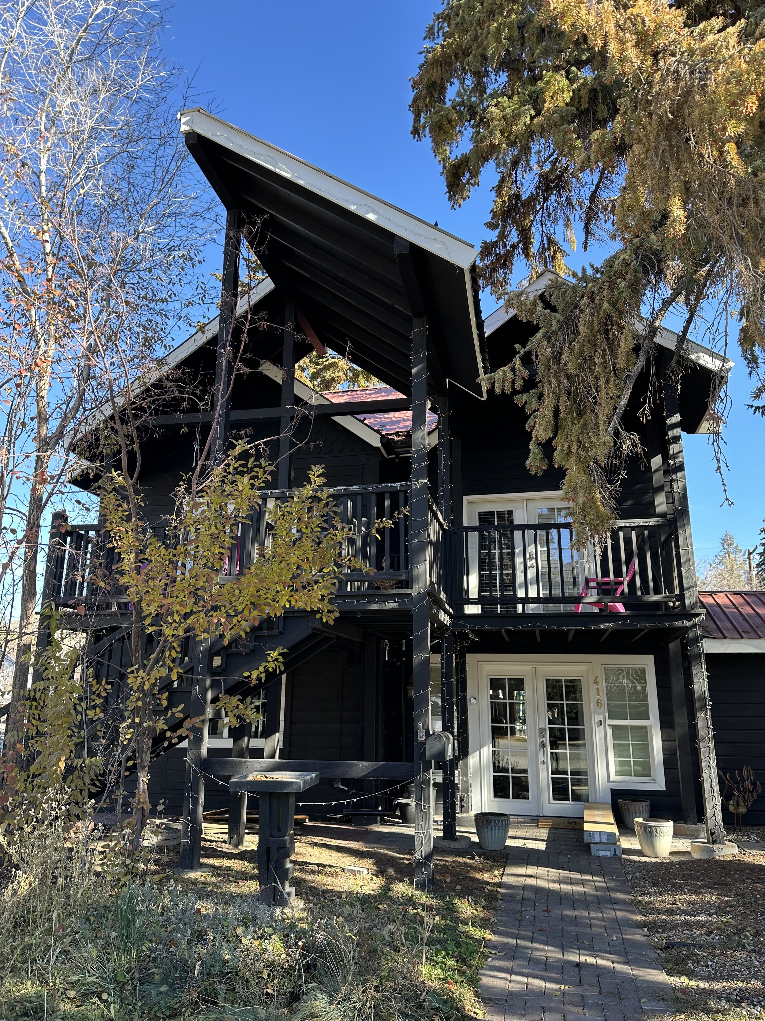 A black two-story house with a balcony and sloped roof, surrounded by trees and plants, with a brick walkway leading to a glass door at the entrance.