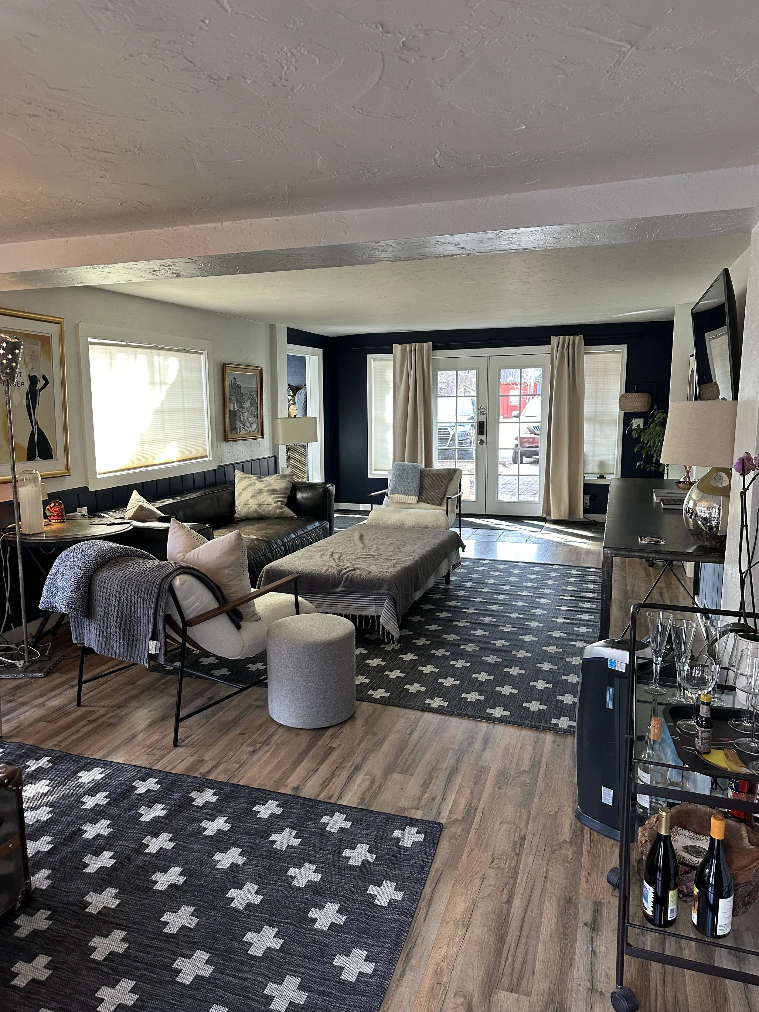 Living room with black sofa, white armchair, patterned rugs, wooden flooring, large windows with blinds, French doors with curtains, side tables with lamps, decorative artwork on walls, and bottles of wine on a cart.