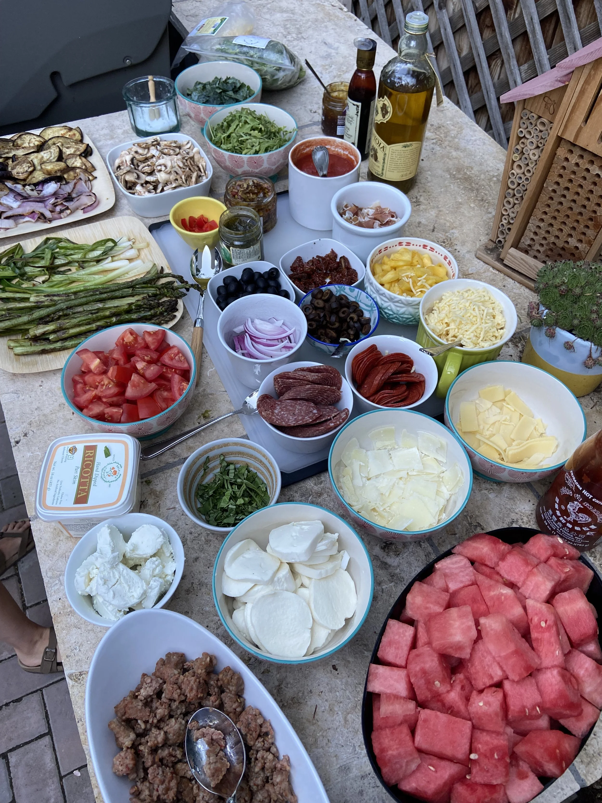 A table with various ingredients for making pizza or similar dishes, including sliced tomatoes, cheese, pepperoni, olives, red onions, cooked ground meat, and various vegetables and sauces.