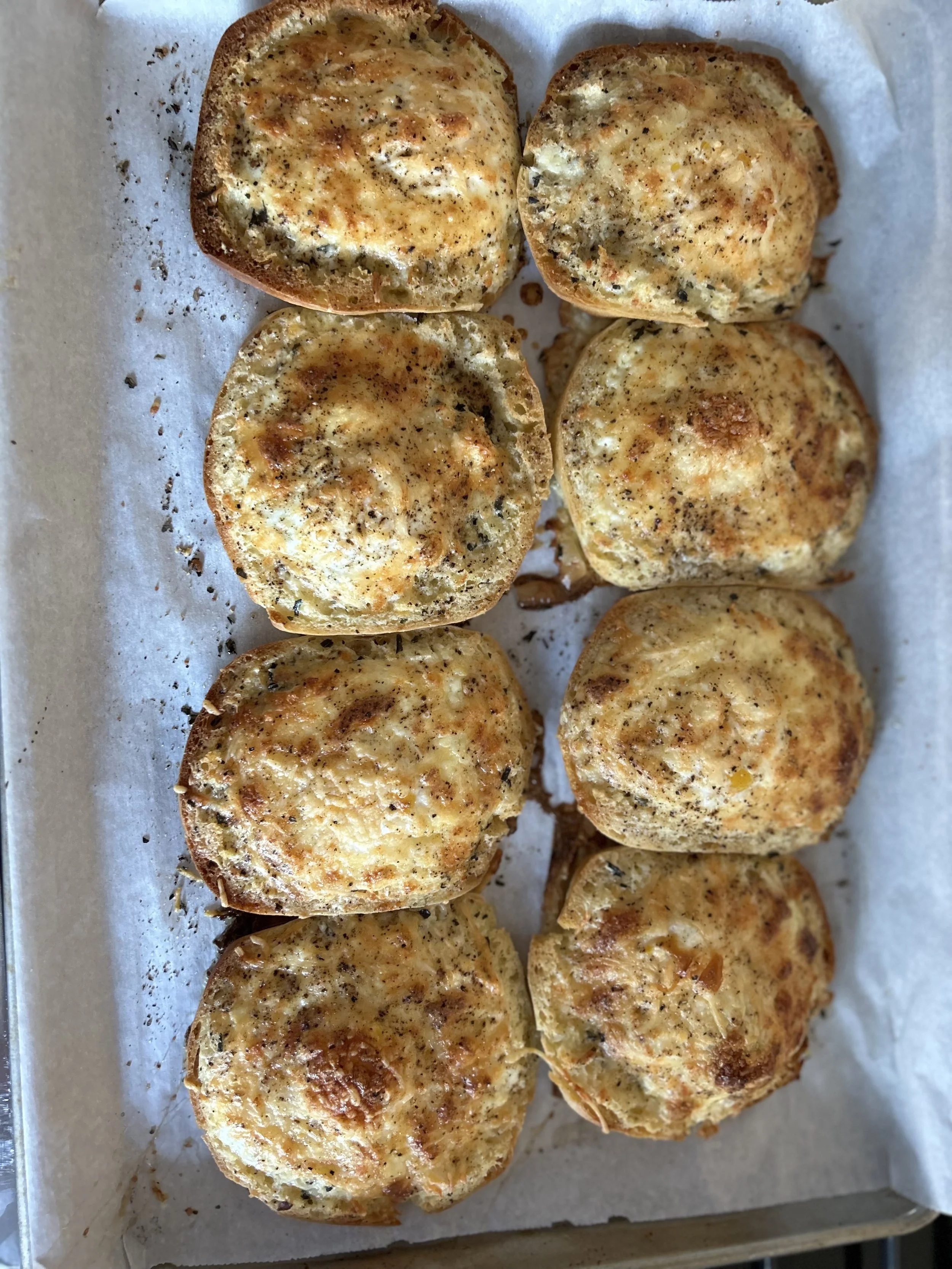 Six garlic bread slices with melted cheese on a parchment-lined baking sheet.