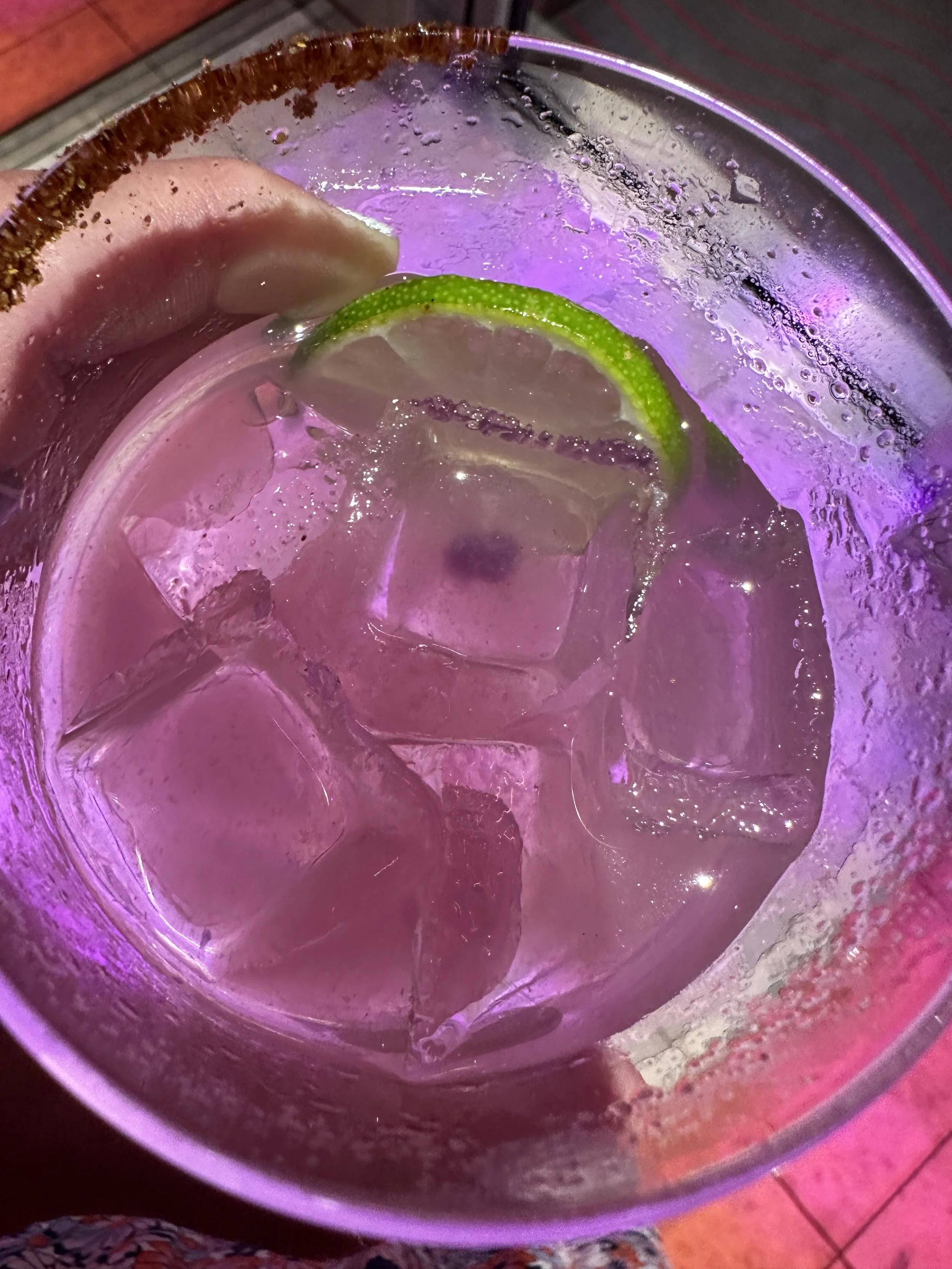 A close-up view of a glass with a lime wedge, ice cubes, and a purple-tinted beverage.