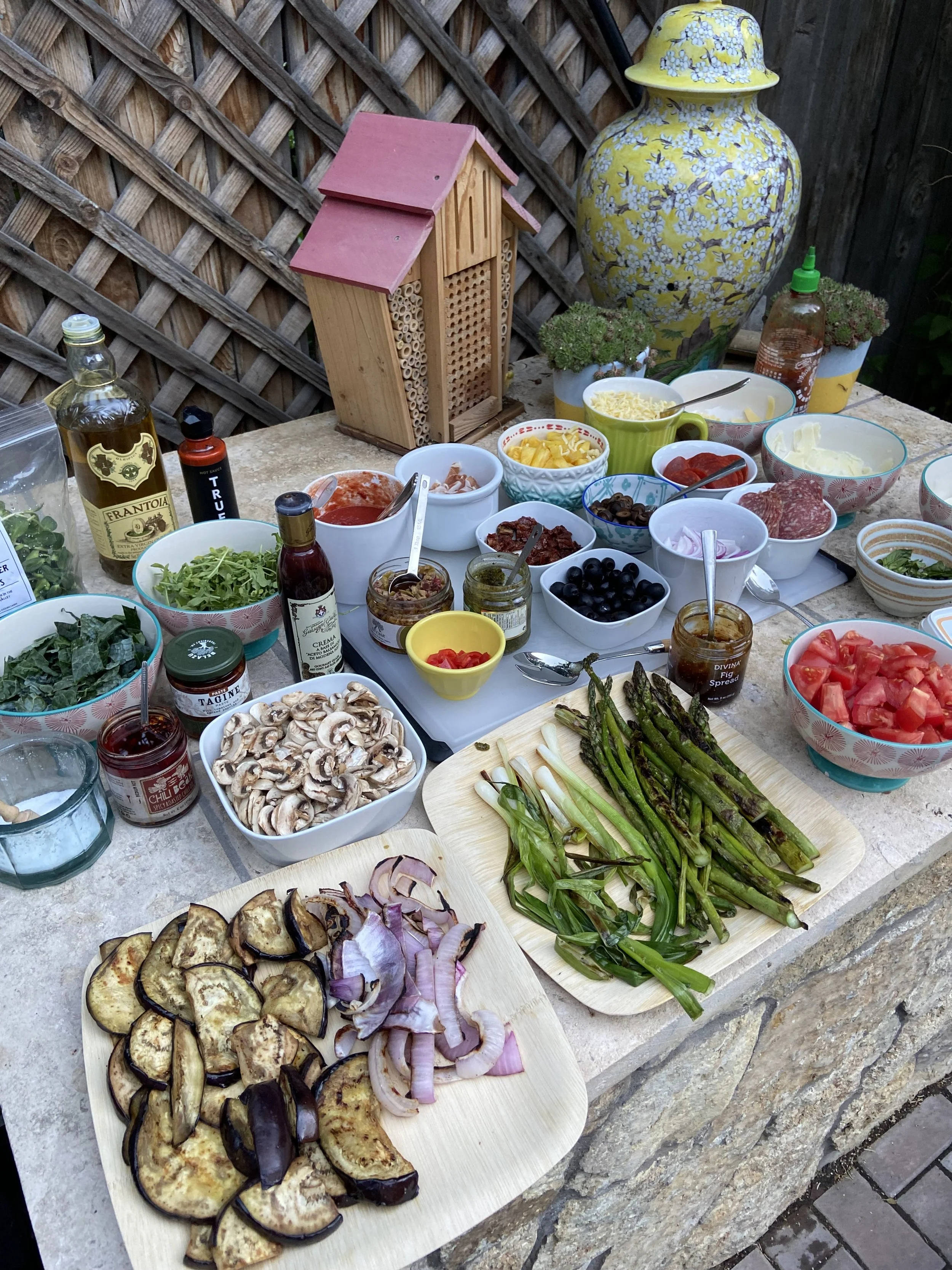 An outdoor table setup for a meal with various vegetables, toppings, condiments, and ingredients, including eggplant, onions, greens, mushrooms, tomatoes, olives, and sauces, with decorative items like a large yellow ceramic urn, plants, and a wooden insect hotel.