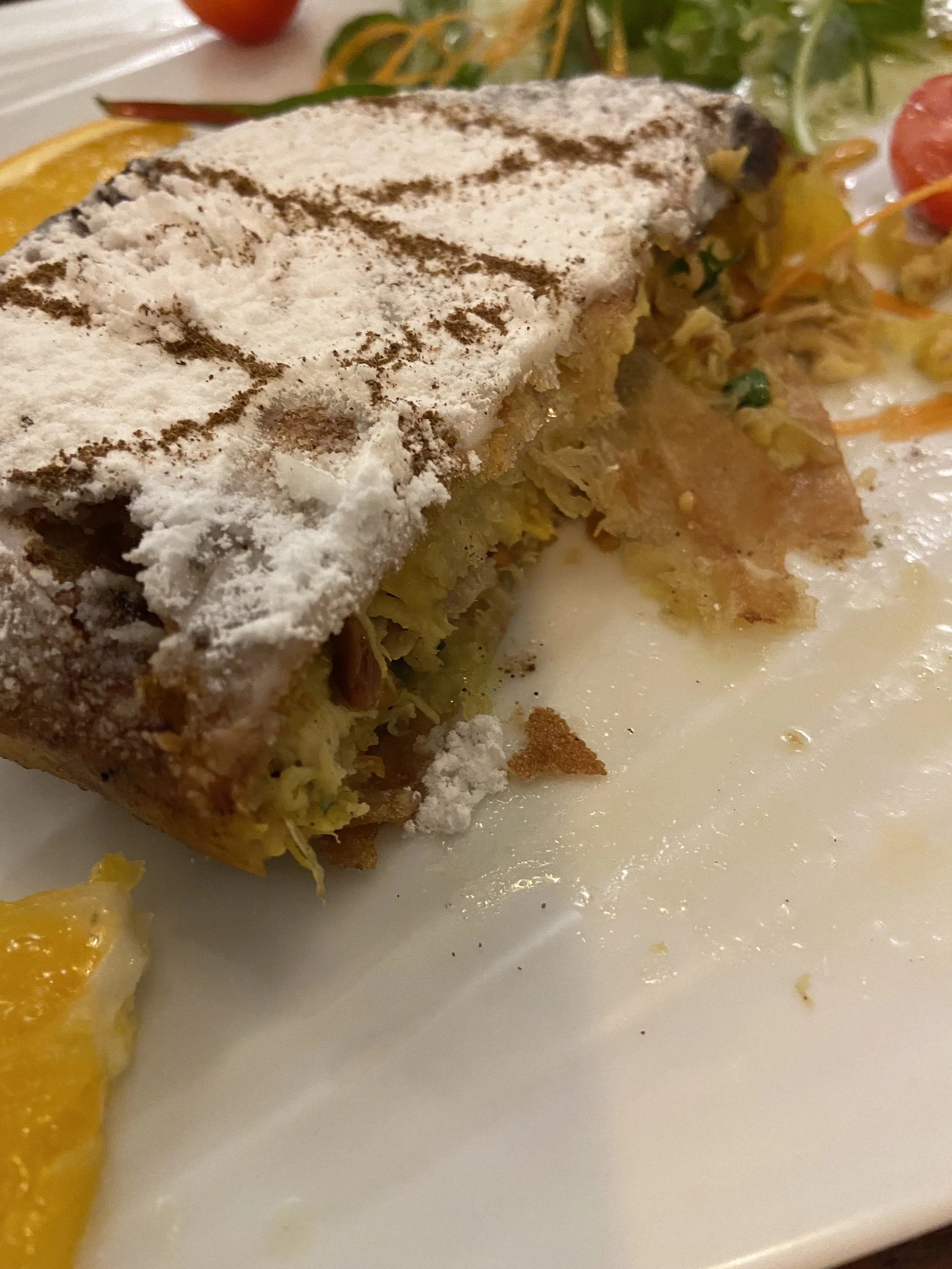 Close-up of a slice of apple strudel with powdered sugar, served with a side of fresh fruit and a sauce or cream on a white plate.