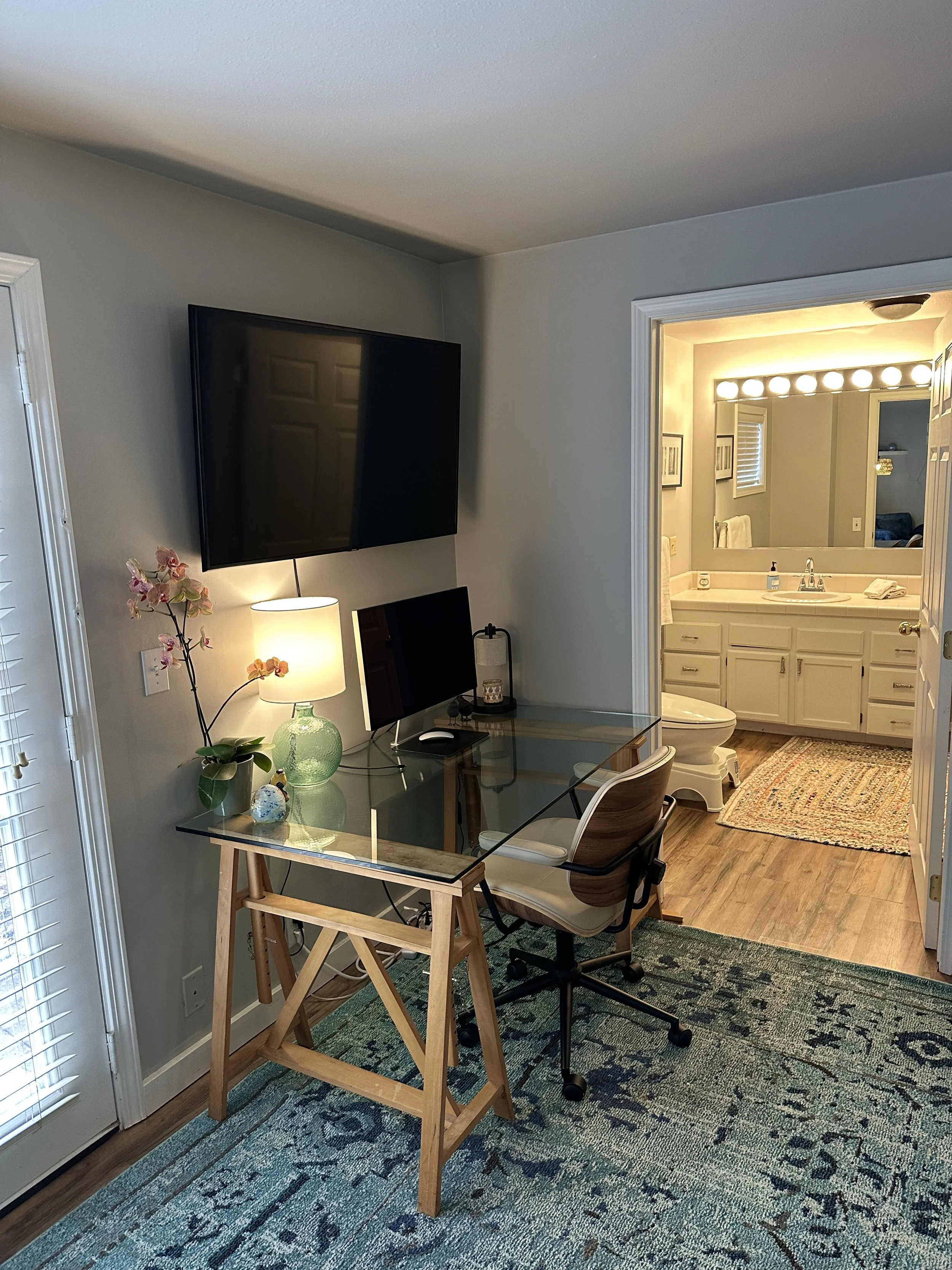 Home office with glass desk, chair, monitors, lamp, orchid plant, and a bathroom visible through an open door in the background.